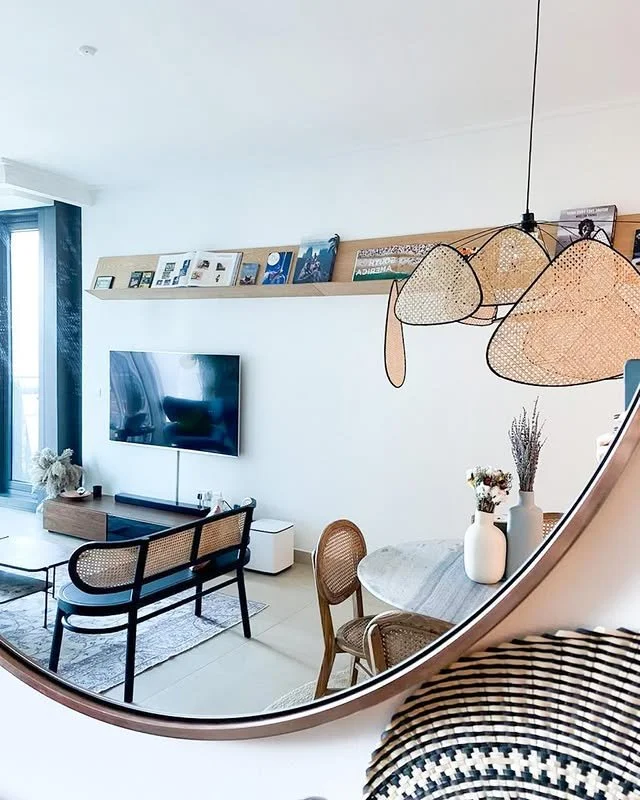 Reflective mirror showing a modern living room with a wall-mounted TV, a wooden shelf with books, a dining table with chairs, and a woven pendant light fixture.