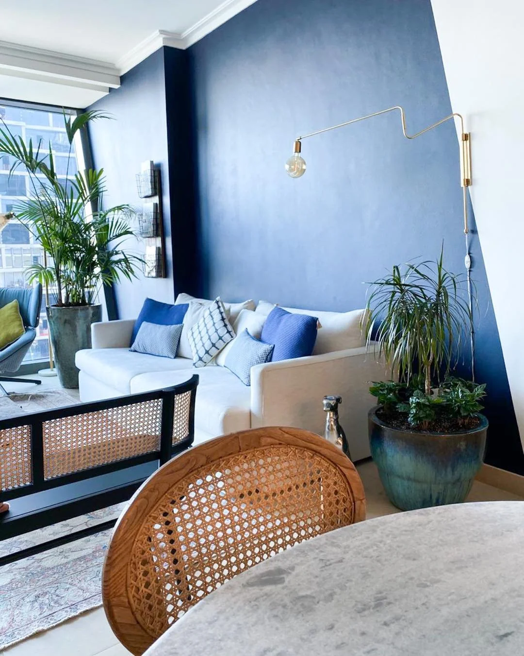 Modern living room with white sofa, blue pillows, large potted plants, wall-mounted light fixture, and large window with city view.