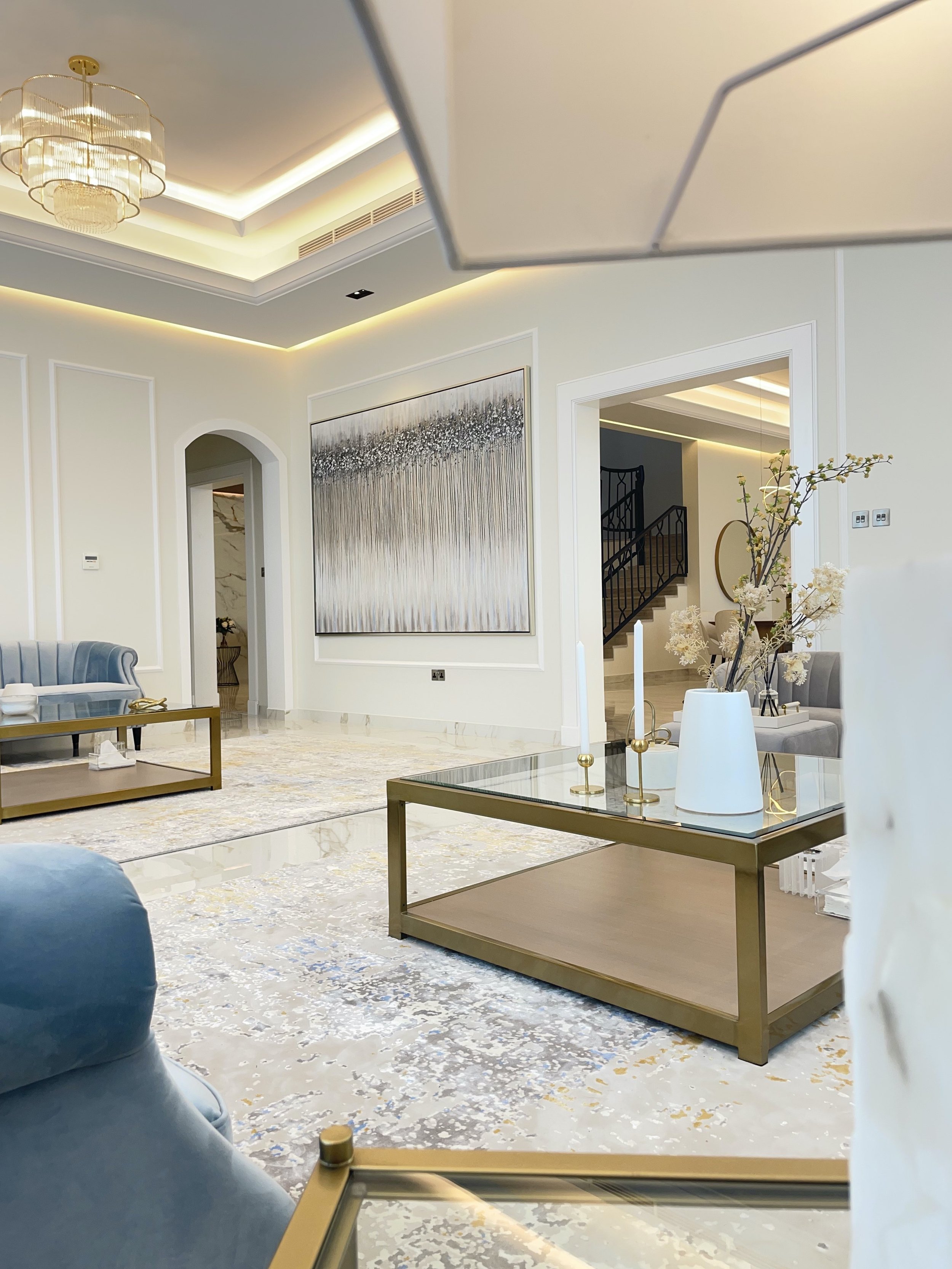 Rasha Ghazaleh | Majlis, Elegant living room with white walls, marble flooring, a large abstract painting, a blue velvet sofa, a glass-top coffee table, and decorative vases with flowers.
