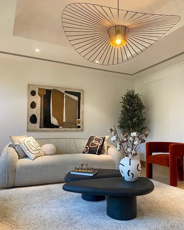 Living room with a beige sofa, abstract wall art, a tall green potted plant, and a modern black coffee table in front, decorated with a white vase holding cotton branches, and a red chair nearby. A unique ceiling light fixture is also visible.