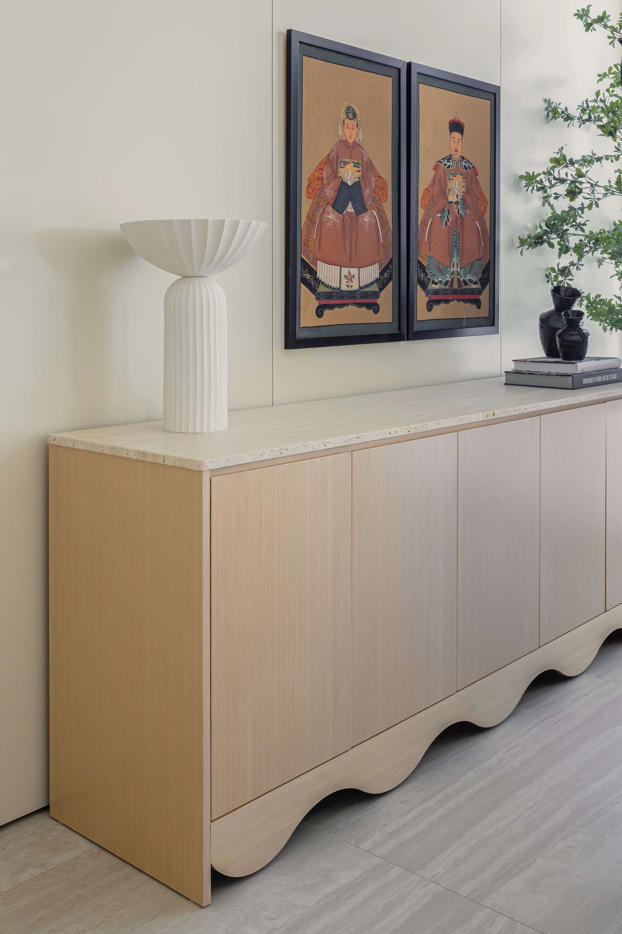 A light-colored sideboard with a wavy bottom edge, decorative white vase with a fluted design on top, and a stack of books with black vases, with framed artwork depicting two traditional Asian figures hanging above, and green foliage on the right.