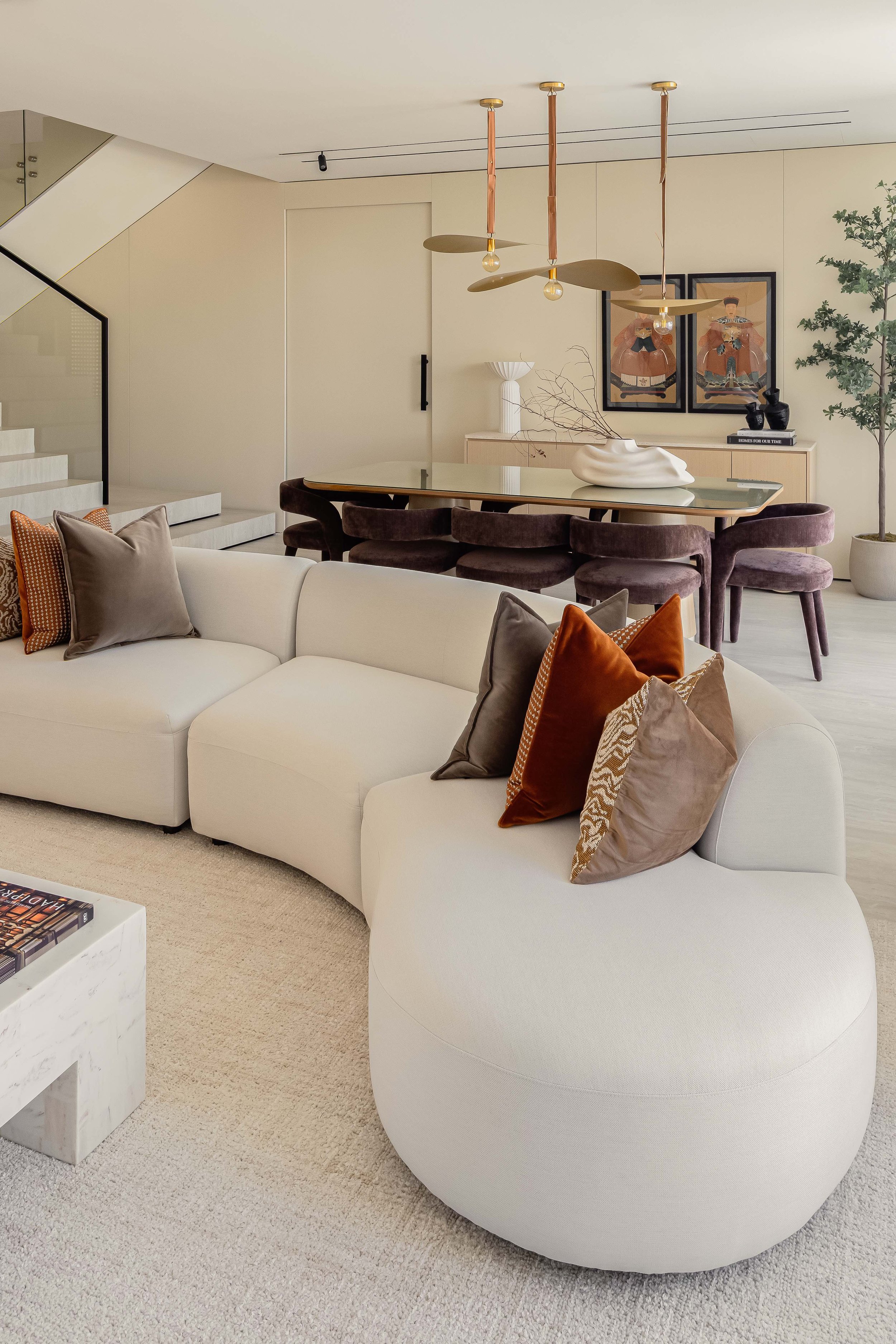 Modern living room with white curved sofa, throw pillows, marble coffee table, beige carpet, dining table with plum velvet chairs, artwork, and a potted plant.