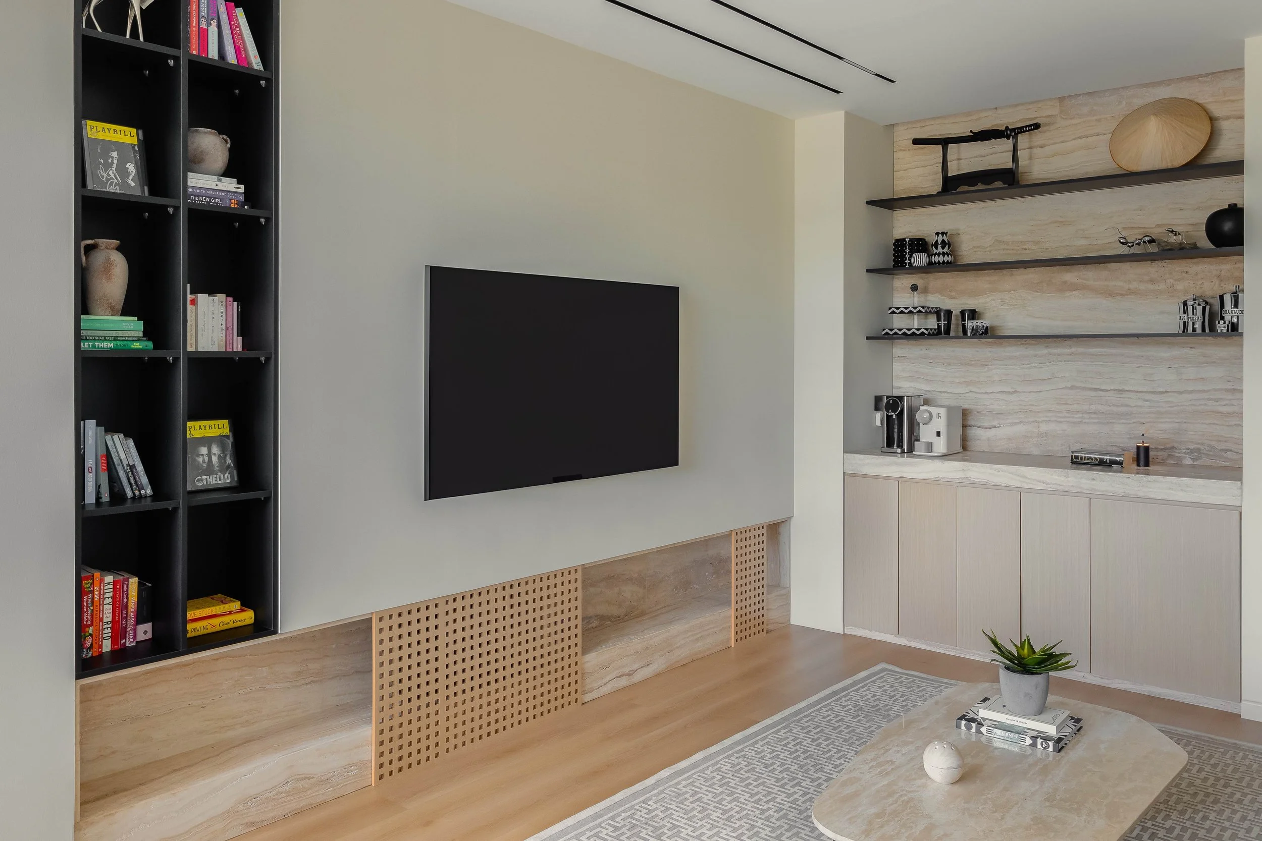 Modern living room with a black bookshelf containing books and decorative items, a wall-mounted flat-screen TV, built-in shelves with decorative objects, a coffee table with a potted plant and magazines, and a light-colored cabinet with coffee-making appliances.
