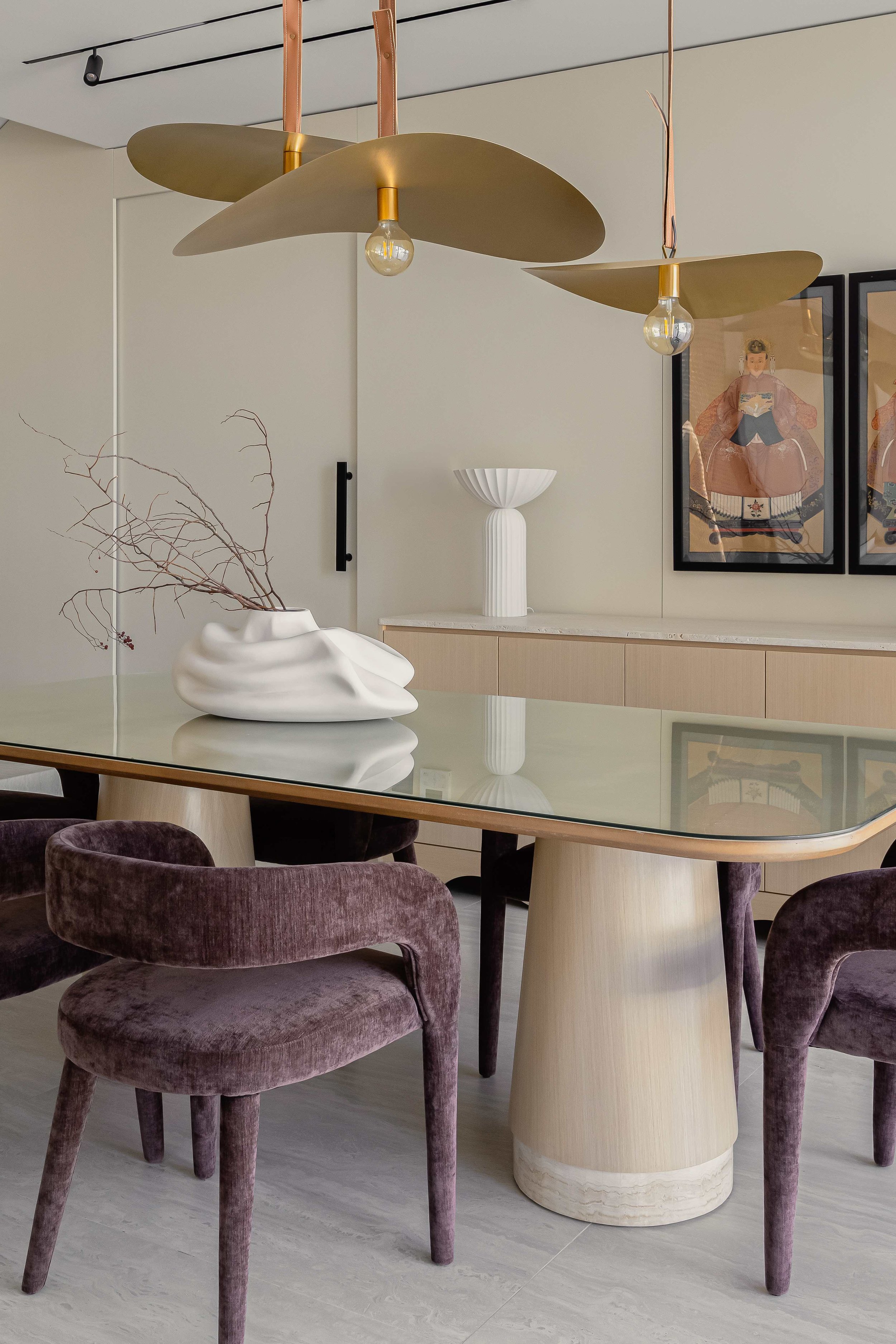 A dining room with a modern chandelier, a glass-top table, purple velvet chairs, and decorative vases and artwork on the wall.