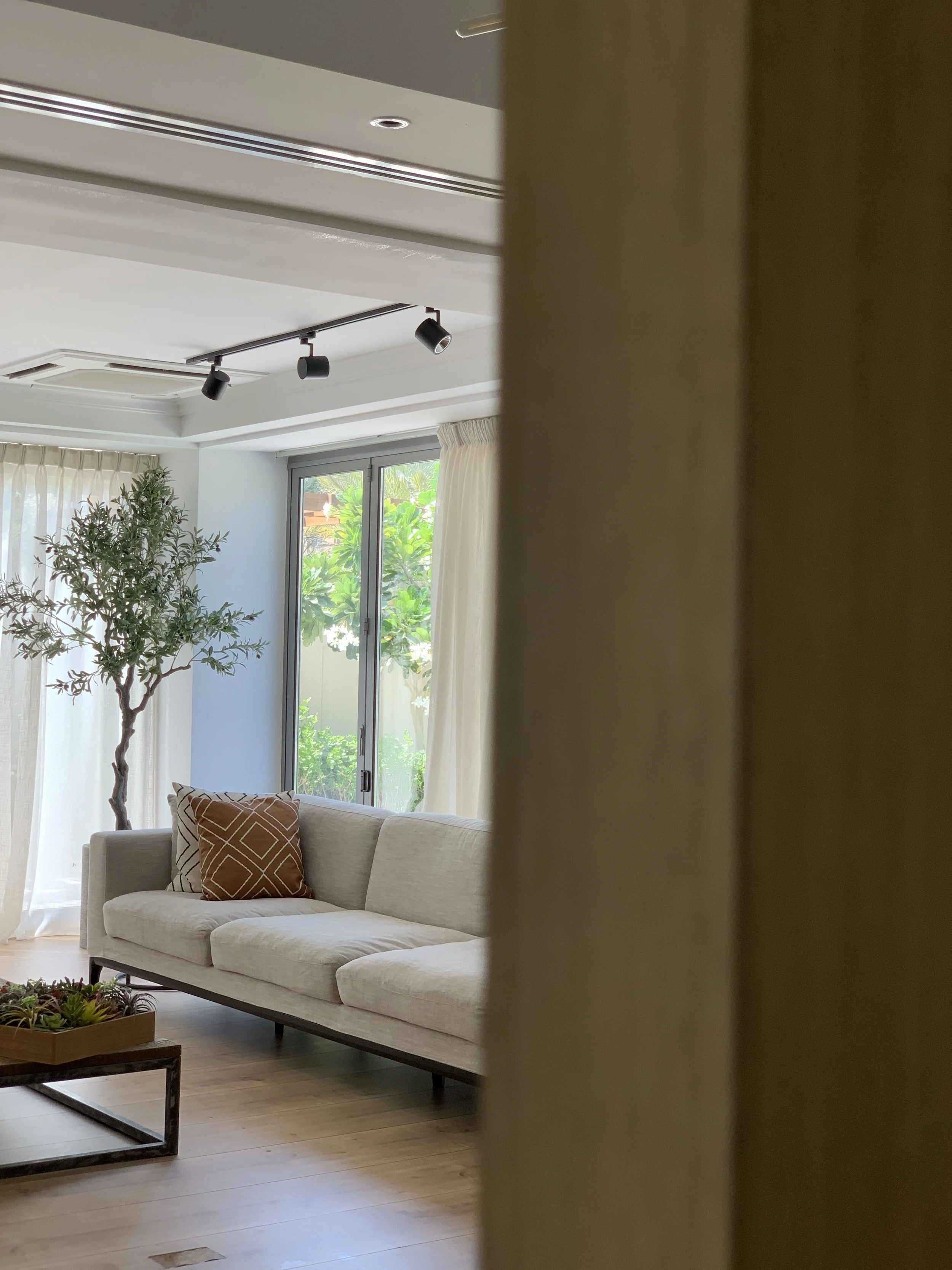 Rasha Ghazaleh | Living room with white couch, decorative pillow, floor plant, large window with cream curtains, wooden flooring, and ceiling lights.
