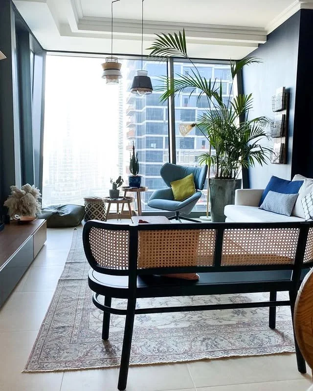 Modern living room with large windows, potted plants, a blue armchair, a white sofa with cushions, a wooden bench, and hanging pendant lights.