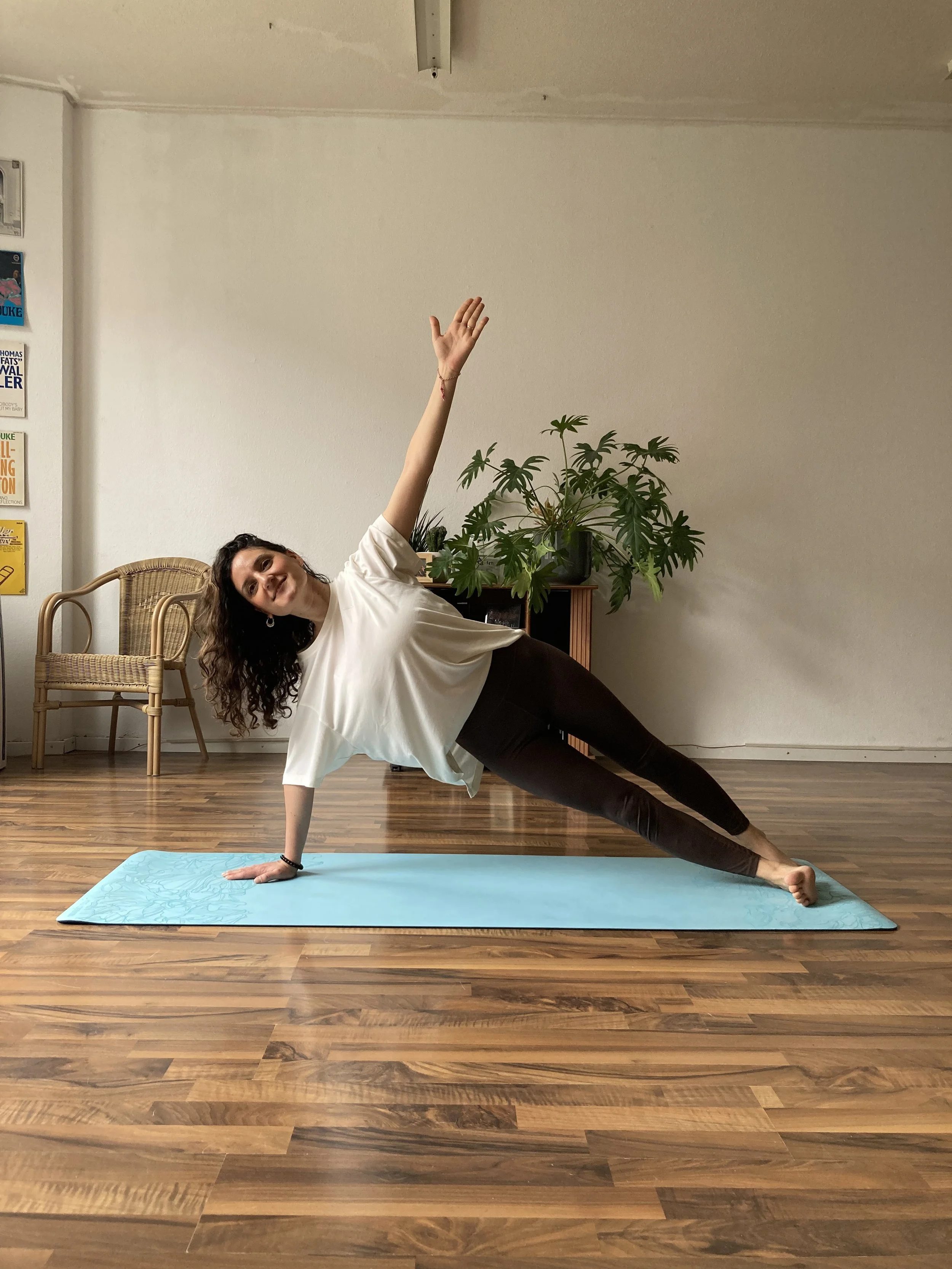 A woman practicing Pilates indoors on a blue mat, in a side plank position with her right hand on the mat and her left arm extended upward. She is smiling and wearing a white t-shirt and dark leggings.