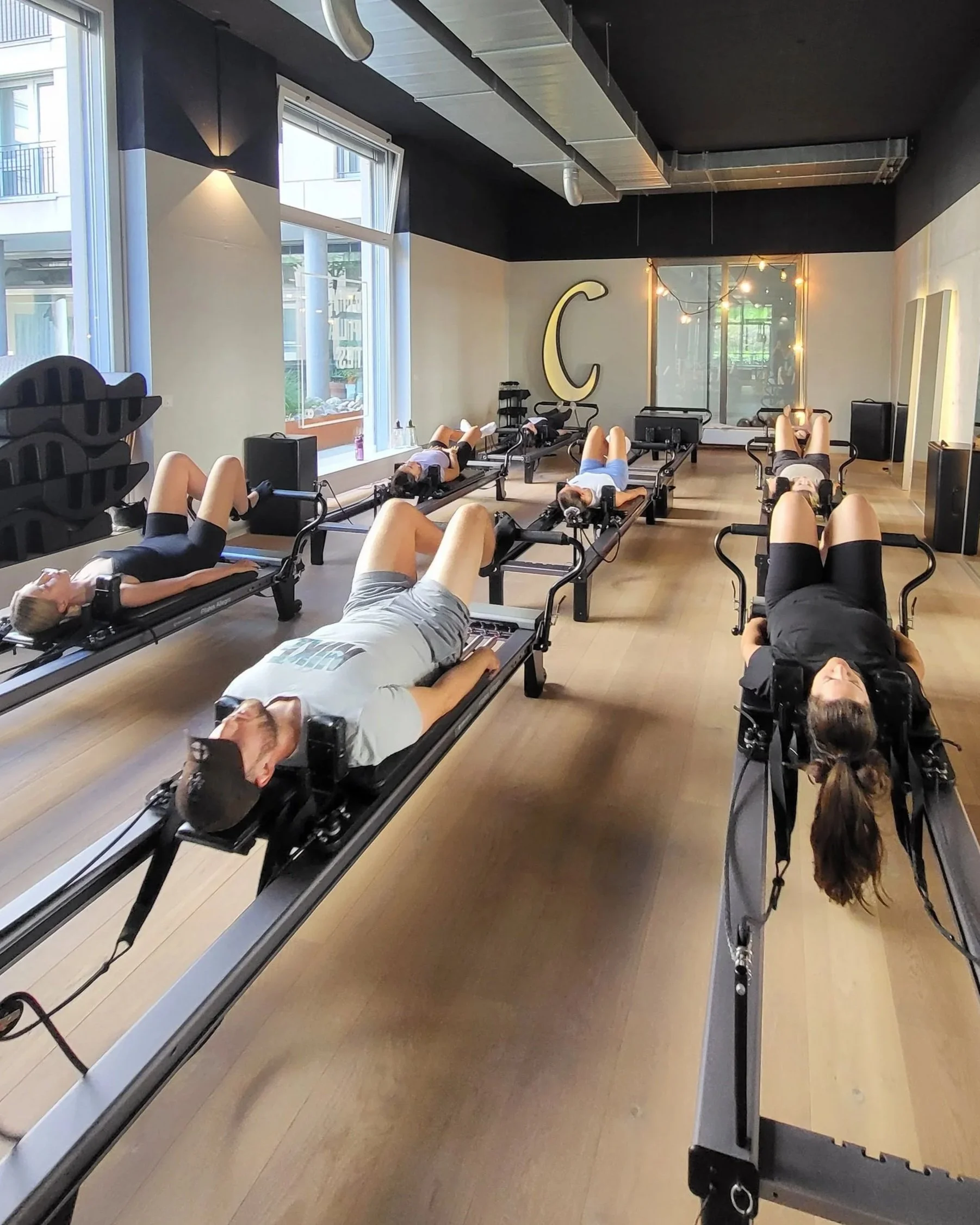 People lying on Pilates reformer machines in a fitness studio with large windows and a decorative letter 'C' on the wall, practicing pilates exercises.