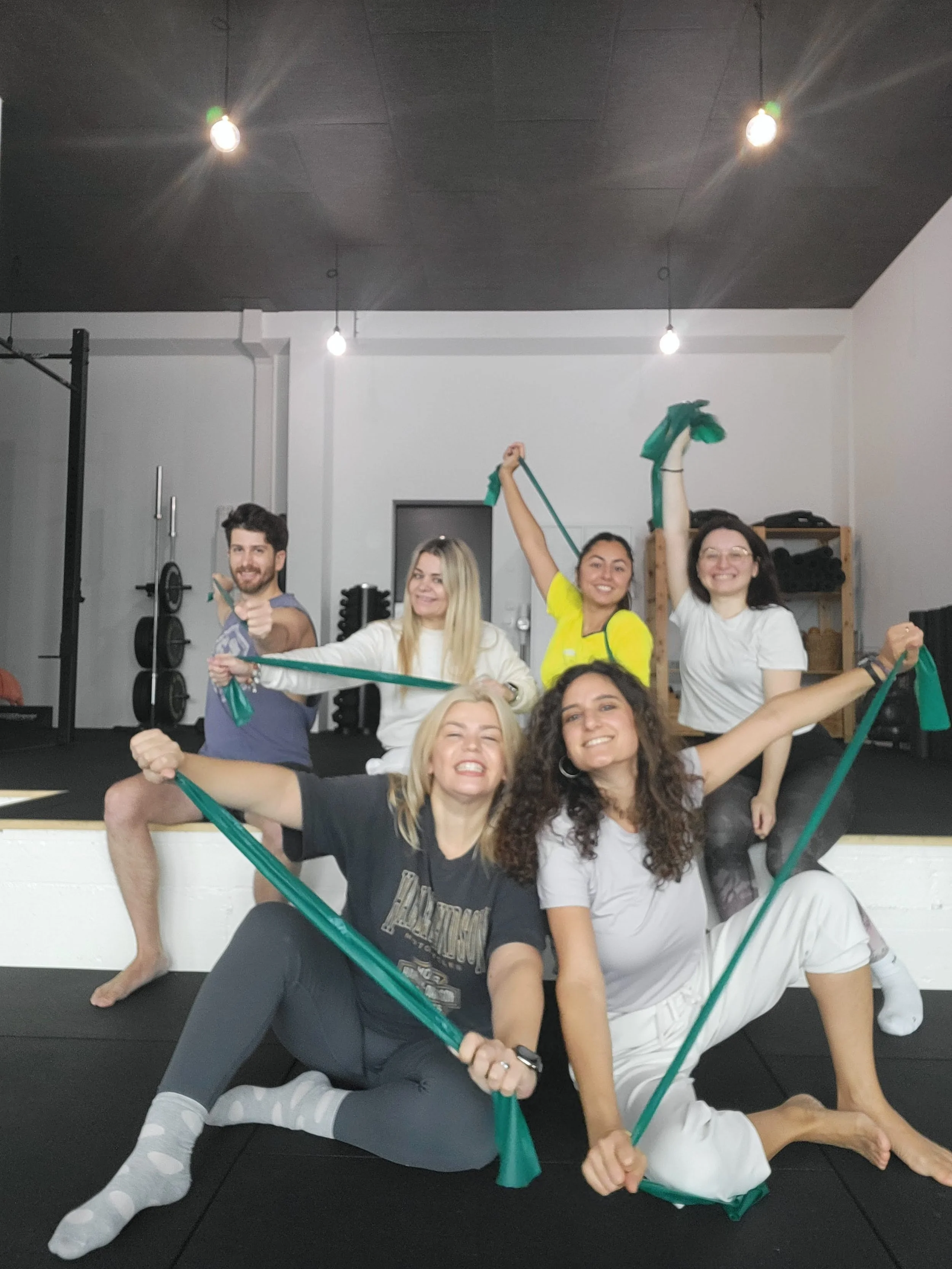 Group of six smiling people in a gym, holding resistance bands, posing enthusiastically for a photo.