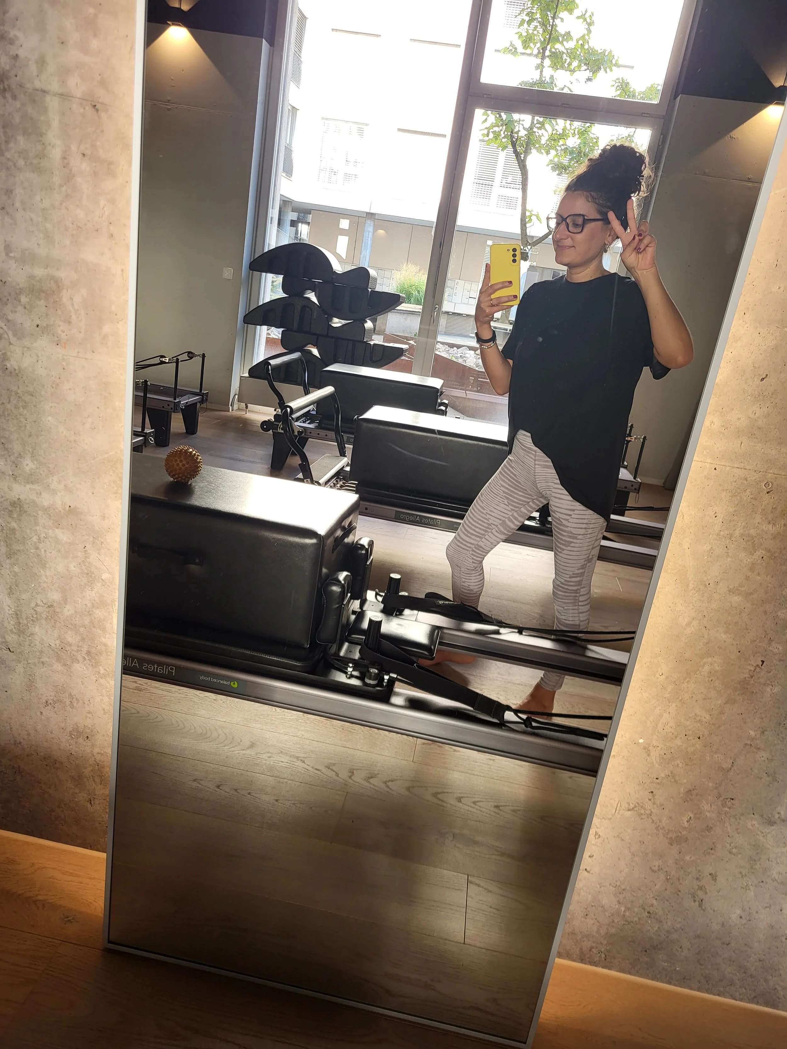 A woman taking a selfie in a gym mirror, making a peace sign with her right hand, wearing glasses, a black t-shirt, and striped leggings.