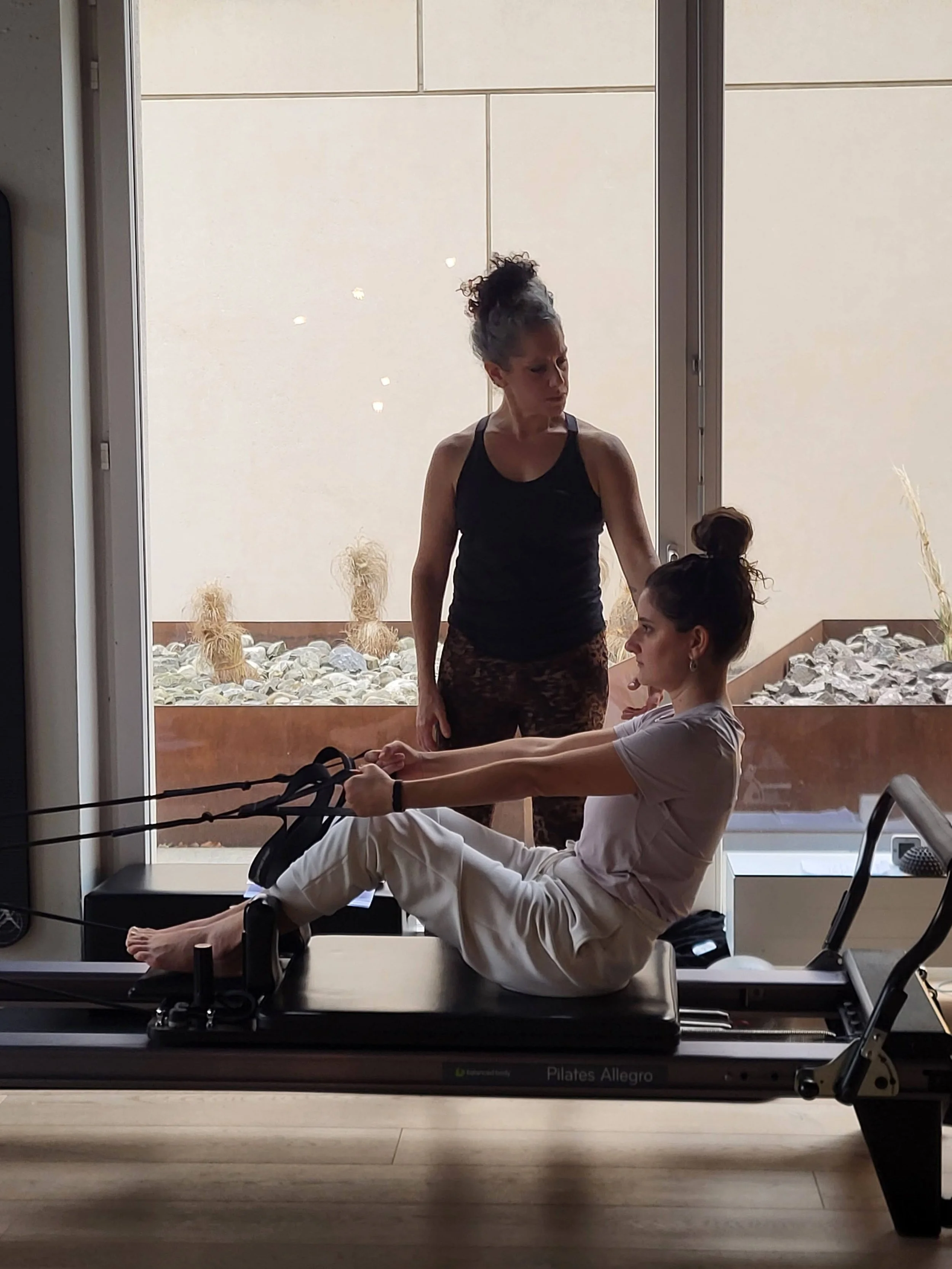A woman is using a Pilates reformer machine for exercise while a trainer guides her, inside a studio with a large window and outdoor decor visible.