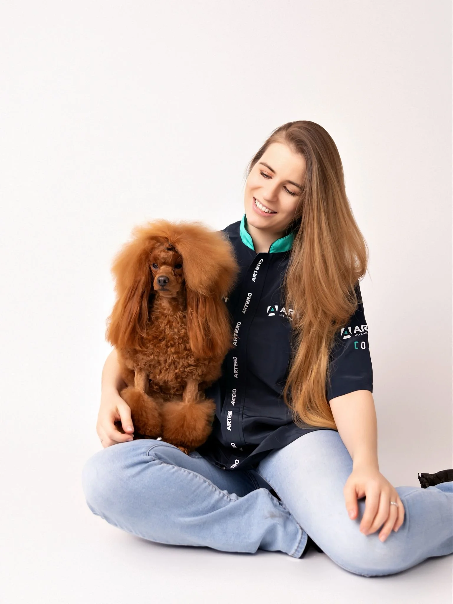 Mujer con cabello largo y rubio, sonriendo, sentada en el suelo, sosteniendo un perro de raza caniche, de pelaje rizado y color marrón, en un fondo blanco.