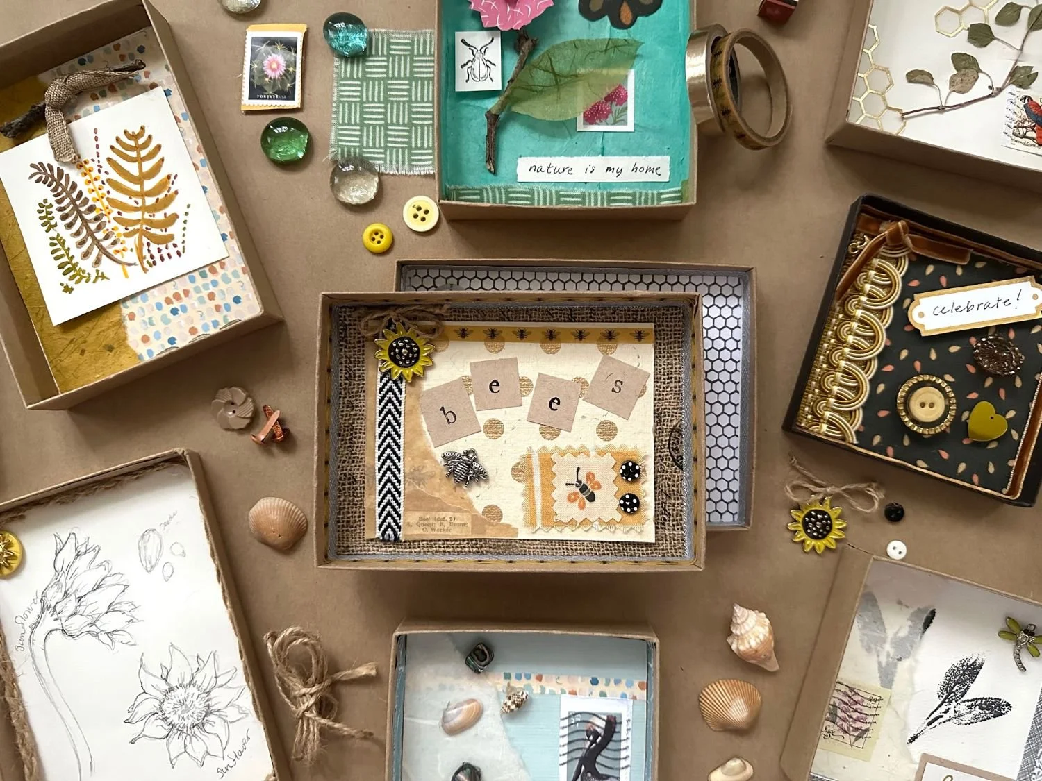 A collection of decorated boxes and craft supplies, including buttons, shell ornaments, illustrated botanical prints, and handmade decorations, arranged on a beige surface.