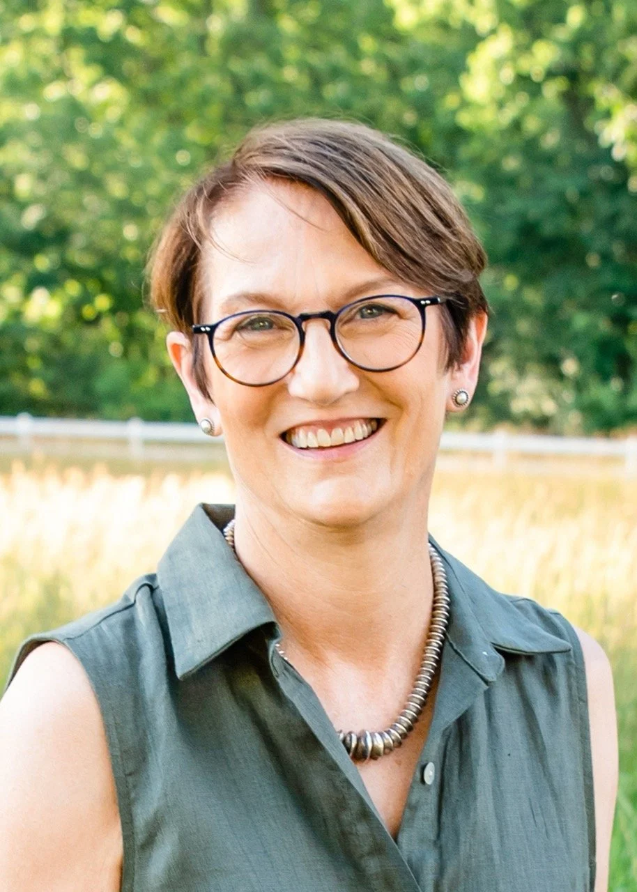 Smiling woman with short brown hair, glasses, and pearl earrings outdoors in a field with green trees in the background.