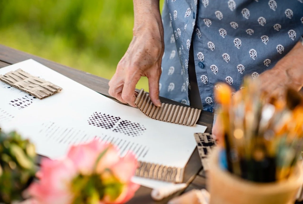 Person arranging cardboard pieces on a table with art supplies and a blurred background of greenery.