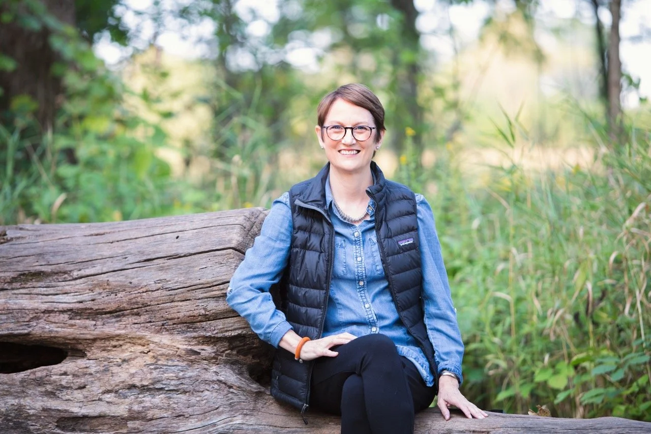 A woman with short hair, glasses, and a denim shirt, smiling while sitting on a large fallen tree trunk in a green forested area.