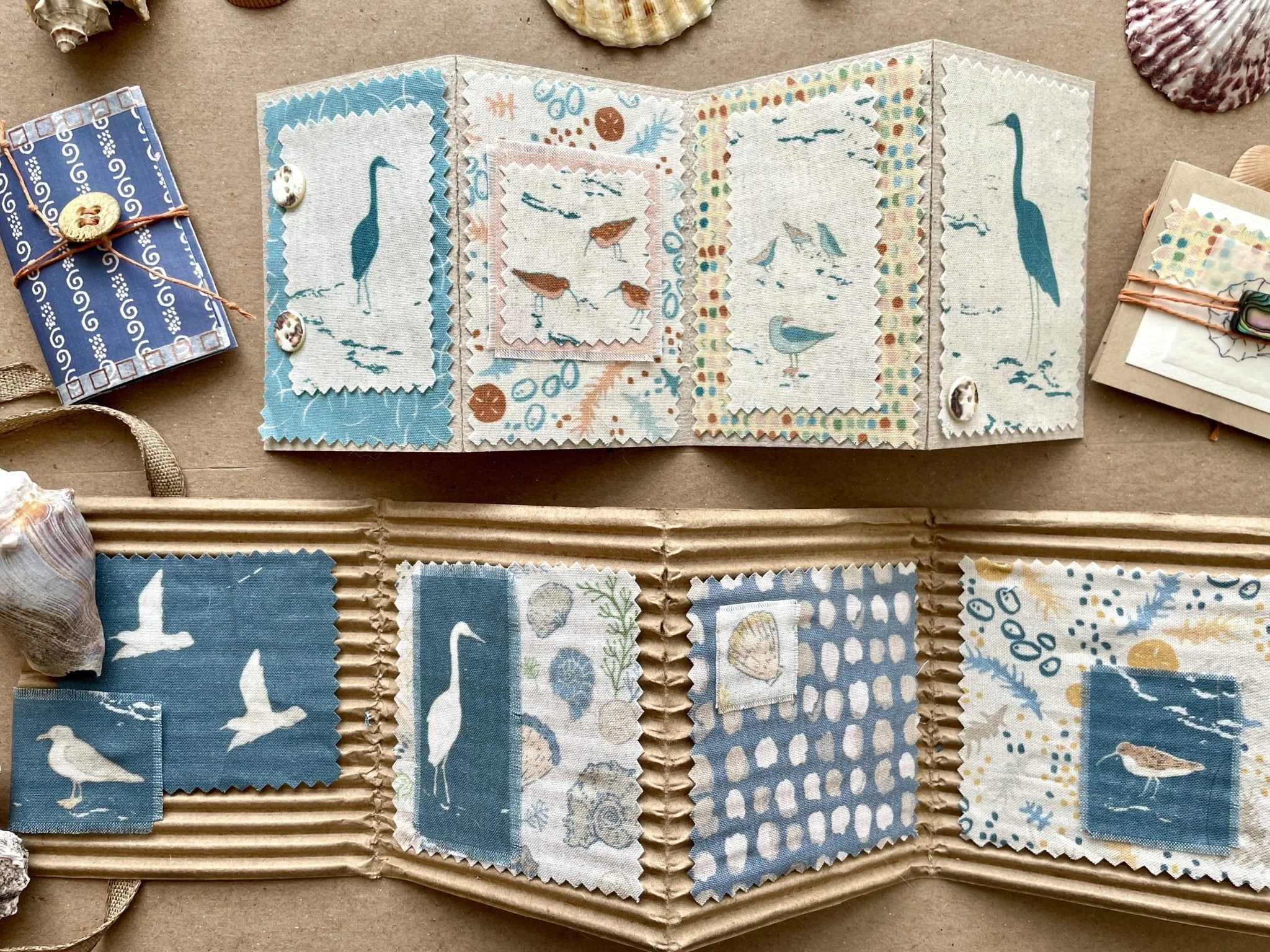 Display of handmade fabric patches with bird and sea-themed designs, arranged on a cardboard surface, surrounded by seashells and small wrapped packages.