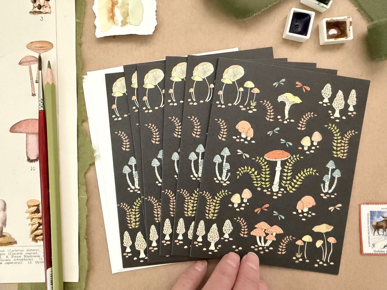 Set of five black sheets of paper with colorful fungus and mushroom illustrations, surrounded by animal and plant illustrations, with art supplies nearby.