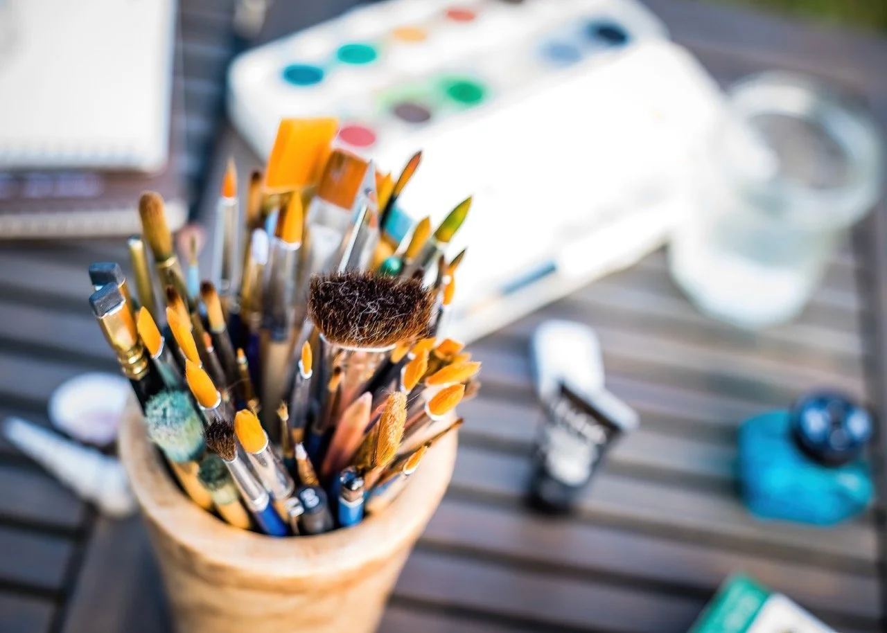 A cup filled with paintbrushes of various sizes and shapes on a wooden surface. Art supplies including watercolor paints, a palette, a small jar, a paint tube, and a digital timer are visible in the background.
