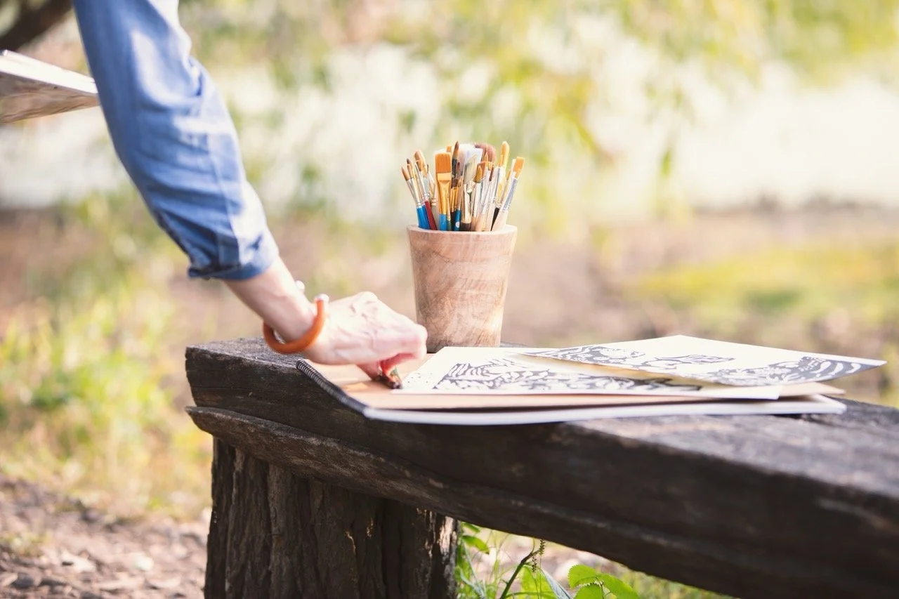 Someone is drawing or writing at a rustic outdoor wooden table with art supplies, including a container of paintbrushes and papers, in a natural setting with blurred greenery in the background.