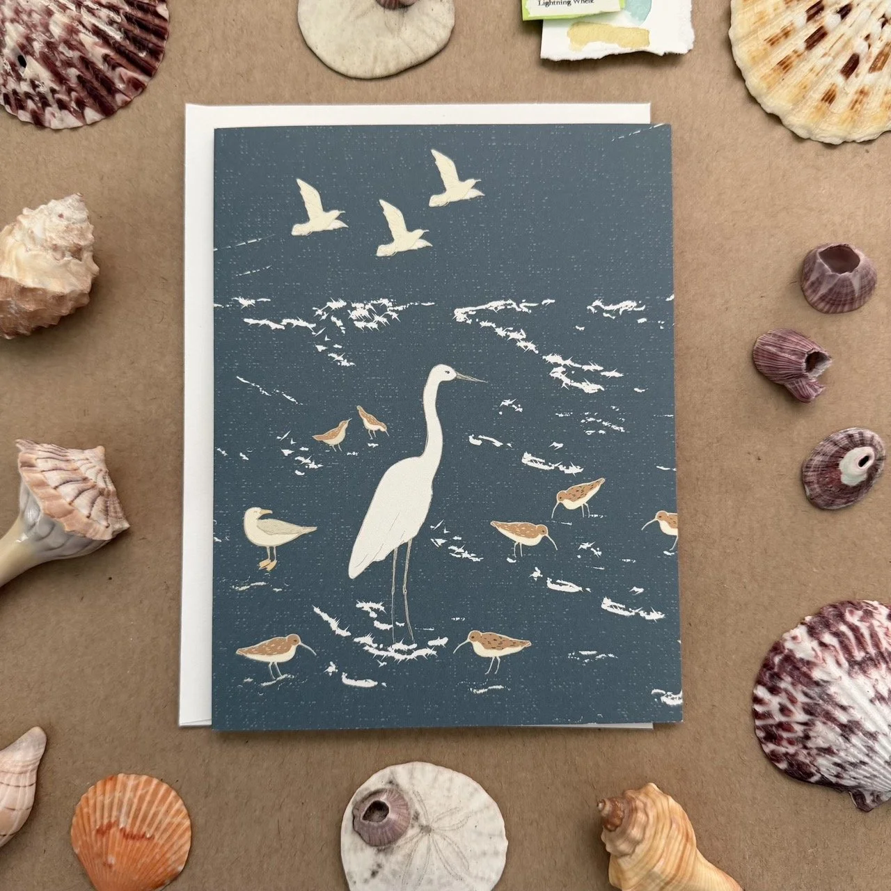 A greeting card with an illustration of a white heron standing in water surrounded by various small birds and flying seagulls, with seashells arranged around it on a tan surface.