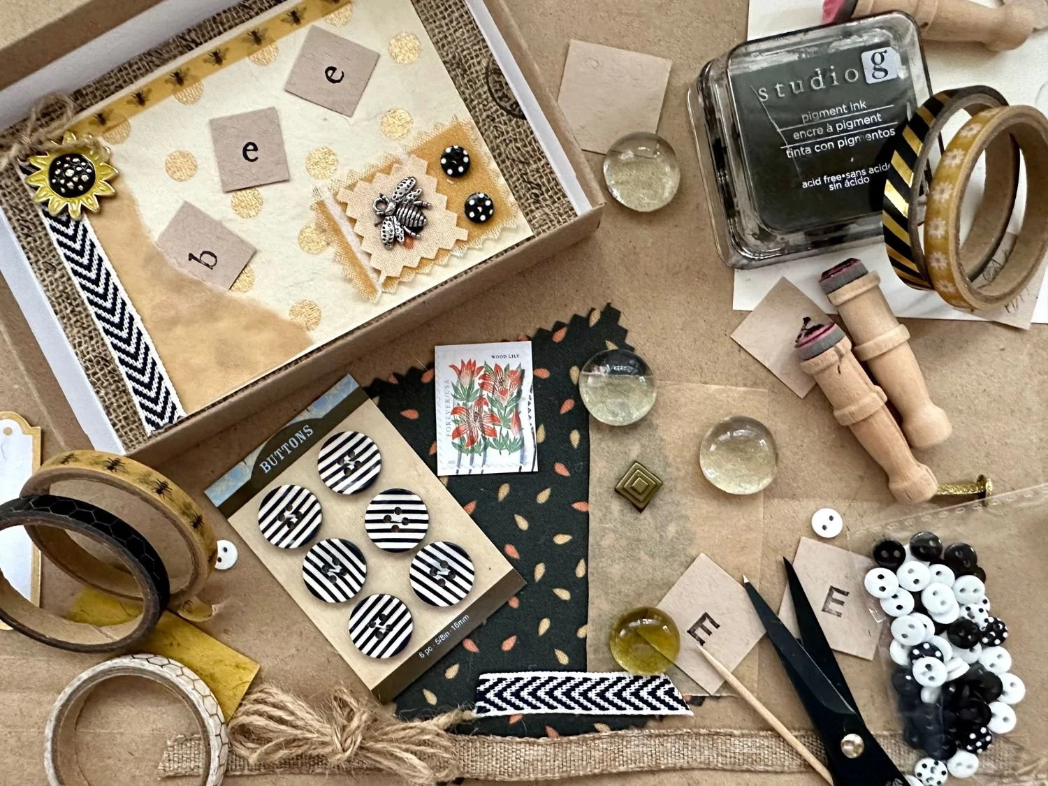 A collection of craft supplies on a table including buttons, washi tapes, stamp ink, decorative papers, a small jar, and various craft tools.