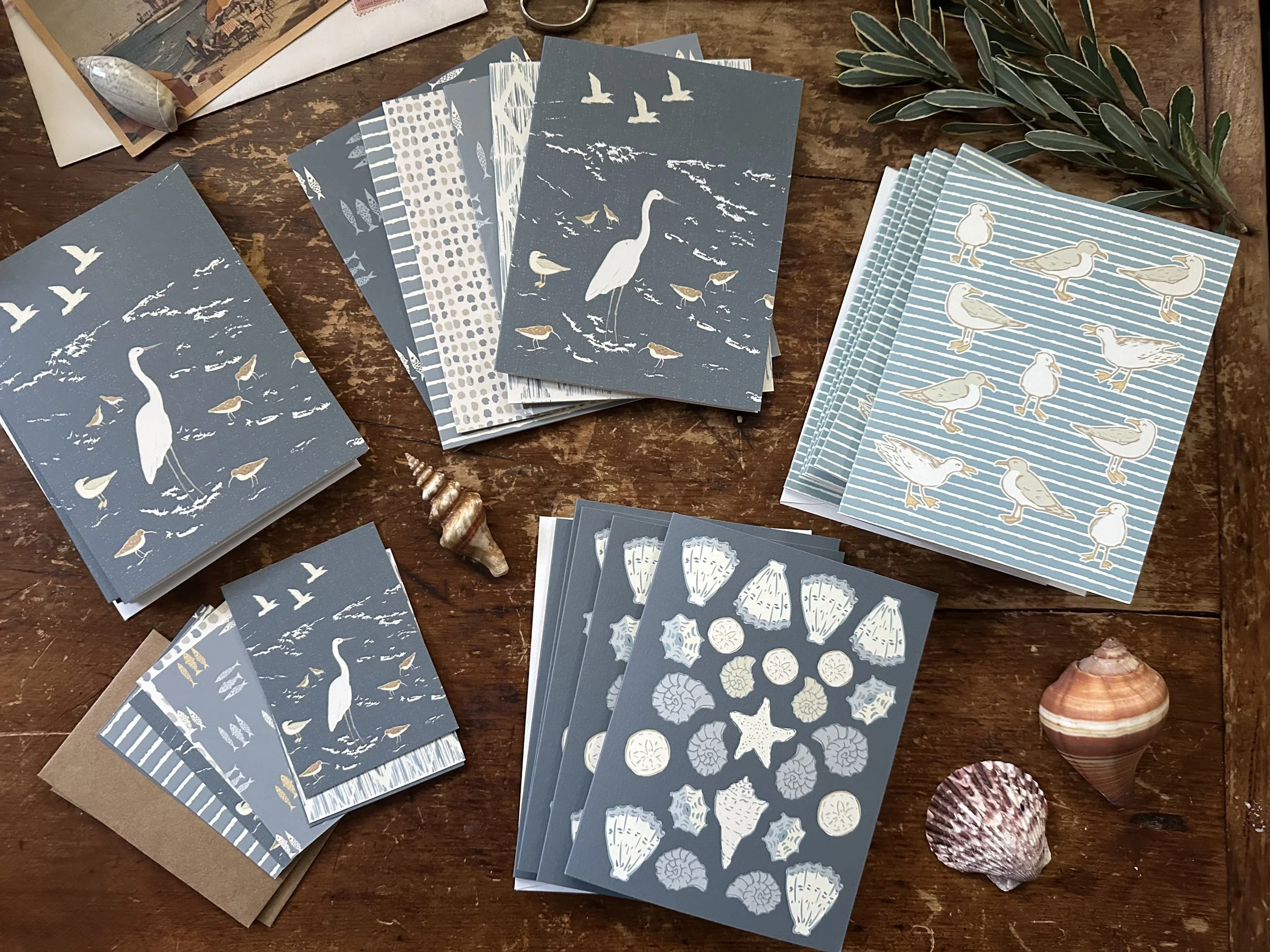 Beige and gray stationery with seaside and bird designs, including shells, starfish, seagulls, and heron, arranged on a wooden table with seashell decorations and green leaves.