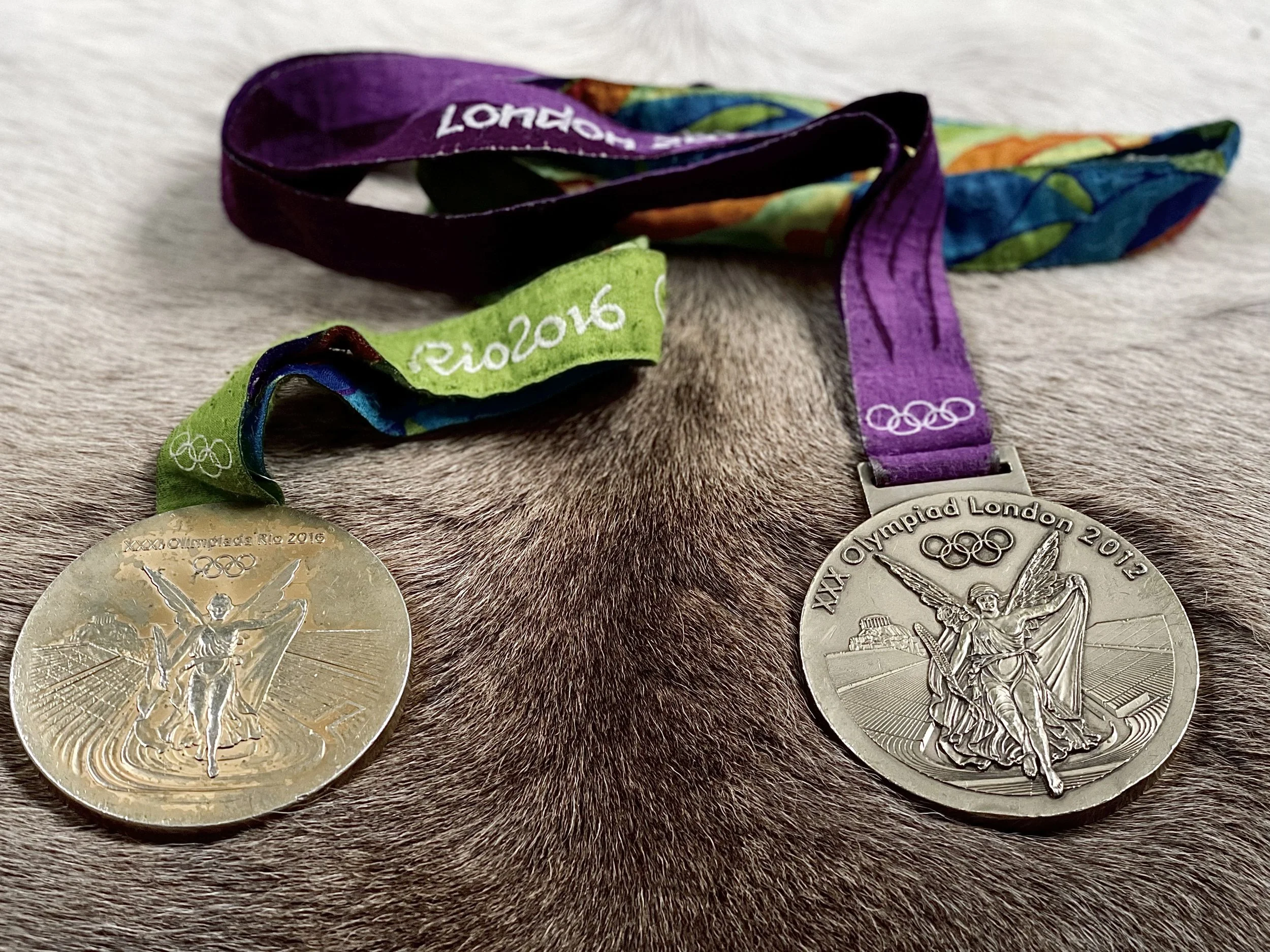 Two Olympic medals from London 2012 and Rio 2016 resting on animal fur, with colorful ribbons attached.