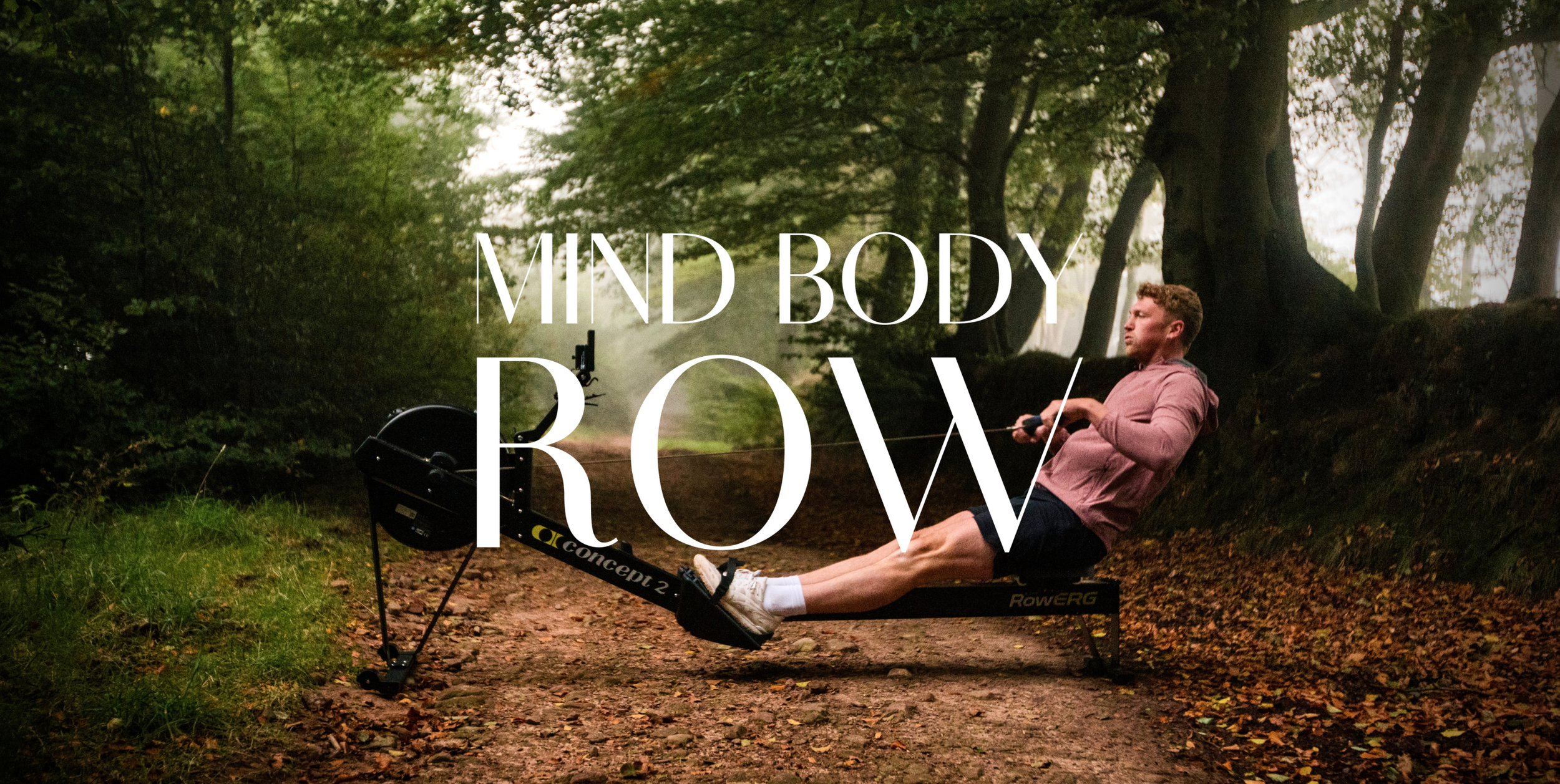 A man exercising on a rowing machine outdoors in a forest surrounded by trees and fallen leaves.