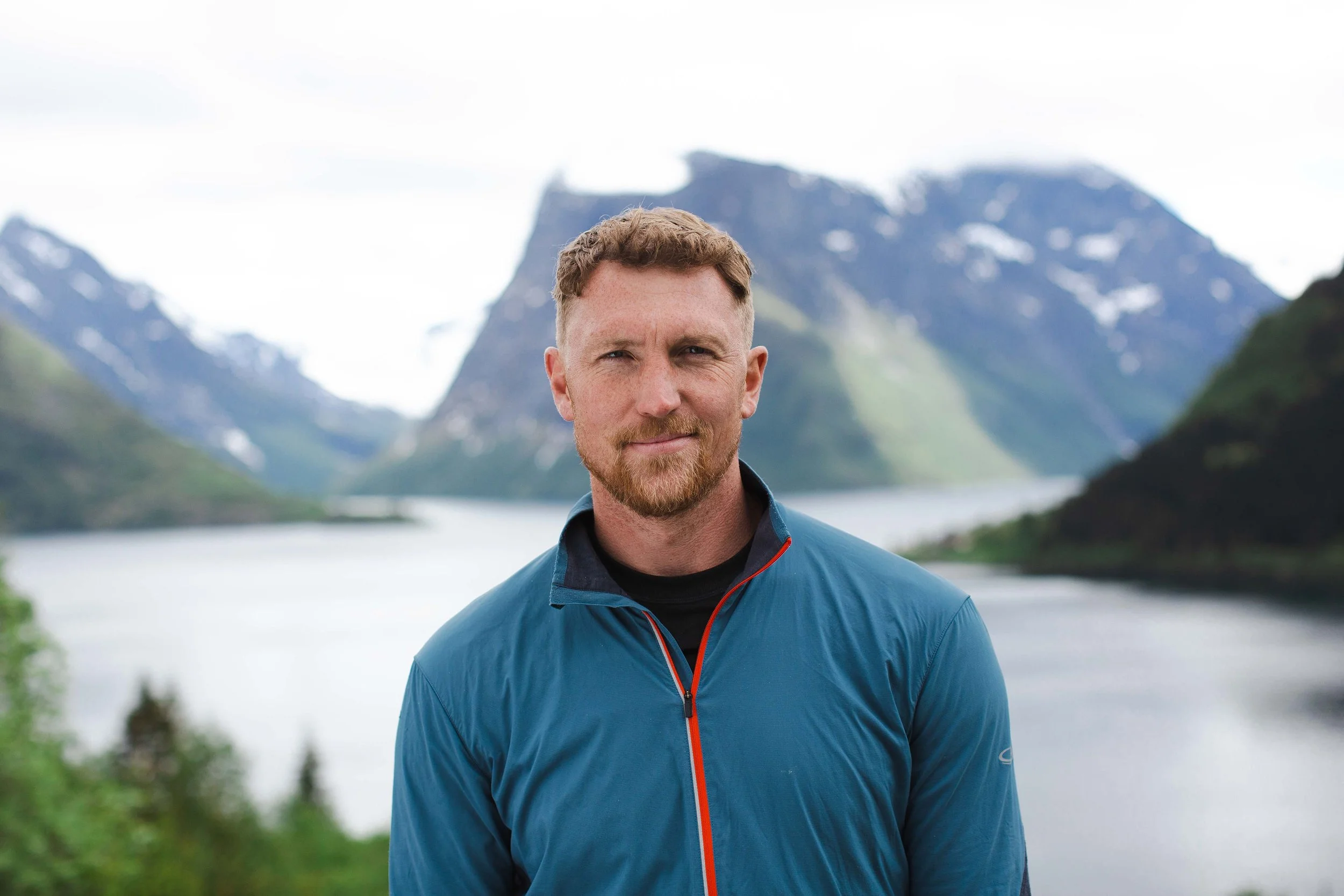 A man with a beard and short hair outdoors with lakes and mountains in the background.