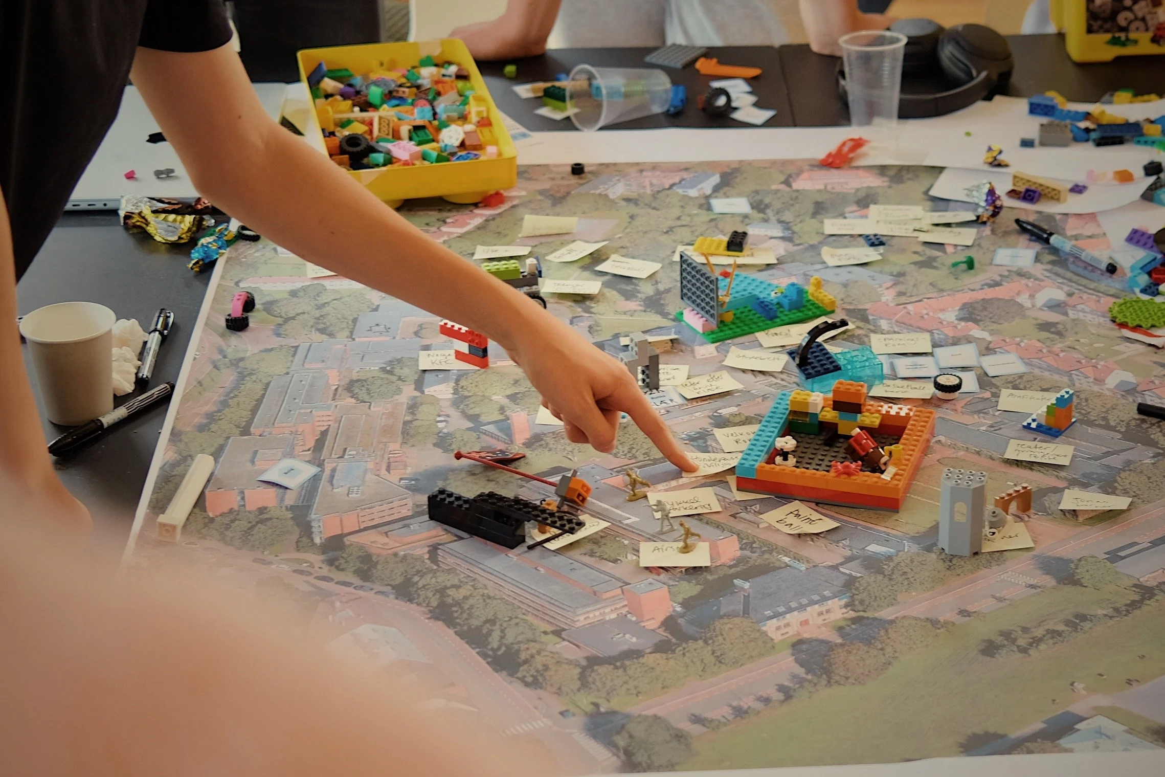 Person som peker på en LEGO-modell på et bord dekket av en stor oversiktskart, med mange små LEGO-figurer, klosser, post-it lapper og verktøy rundt.