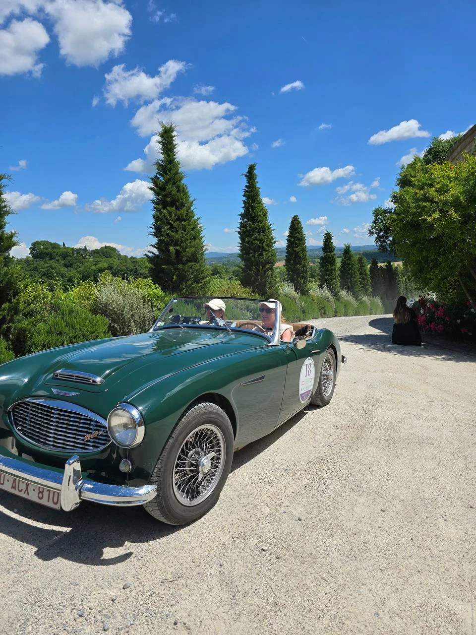 A vintage green convertible car with two women sitting inside, parked on a gravel road with tall trees and a blue sky with white clouds in the background.