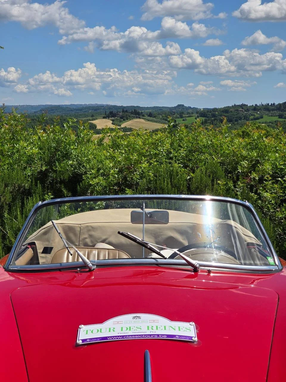 A red vintage convertible car with a beige interior parked in a countryside landscape with green fields, bushes, hills, and a sky filled with fluffy clouds.