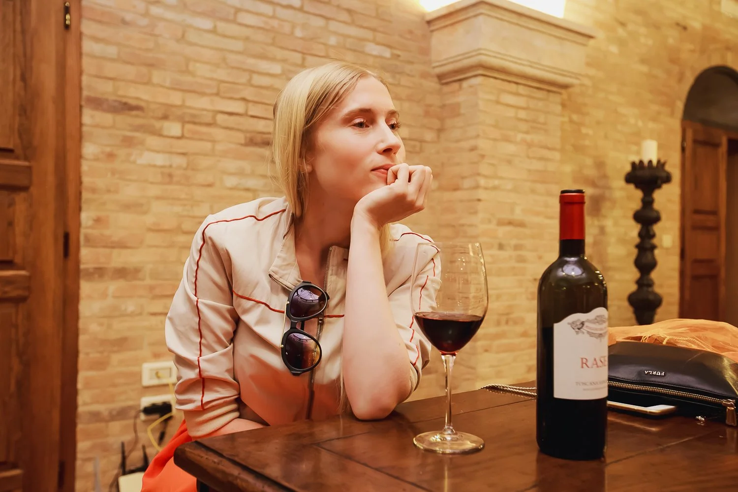 A woman sitting at a wooden table with a glass of red wine and a bottle of wine, resting her chin on her hand, in a room with brick walls.