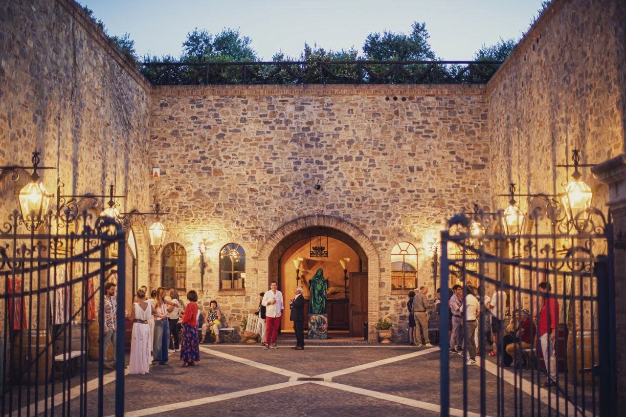People gathered in a courtyard with stone walls and lantern lights, near a large arched entrance, during evening.
