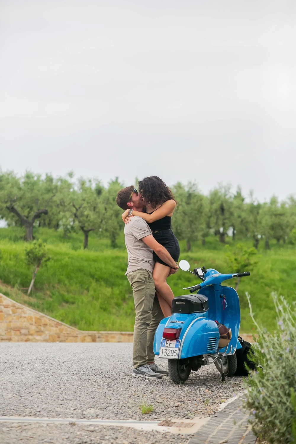A couple is kissing outdoors next to a blue scooter on a stone-paved area with green trees in the background.
