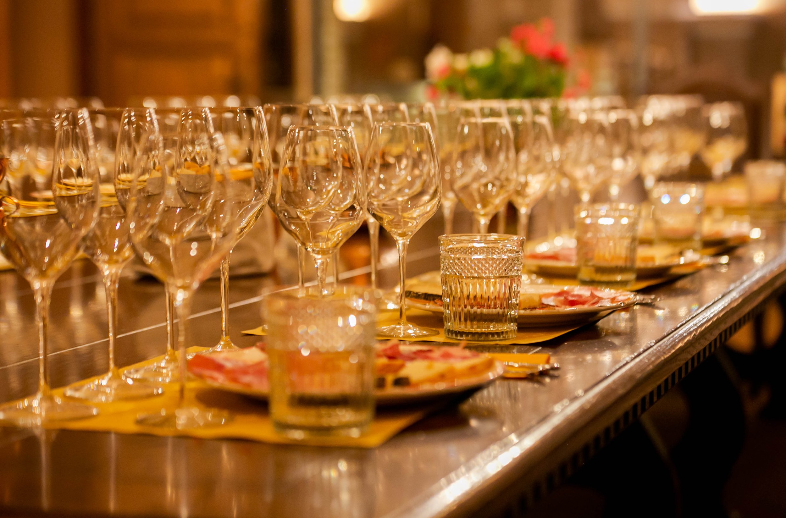A long table set for a celebration with empty wine glasses, small water glasses, plates of finger foods, and floral centerpiece in the background.