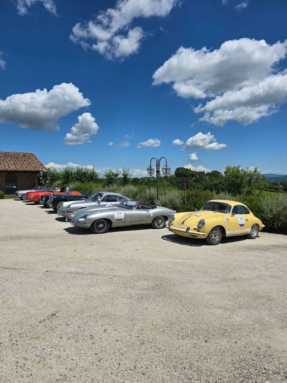 A lineup of vintage race cars parked on a gravel lot under a bright blue sky with fluffy clouds, greenery in the background, and a stone building to the left.