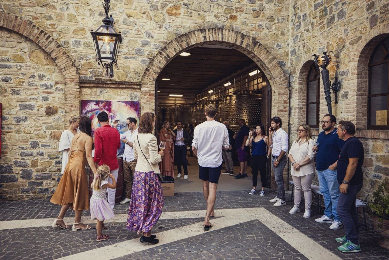 People gathered outside a winery or brewery entrance, socializing, some holding wine glasses, with a stone wall, arched doorway, artwork, and illuminated tanks inside visible.