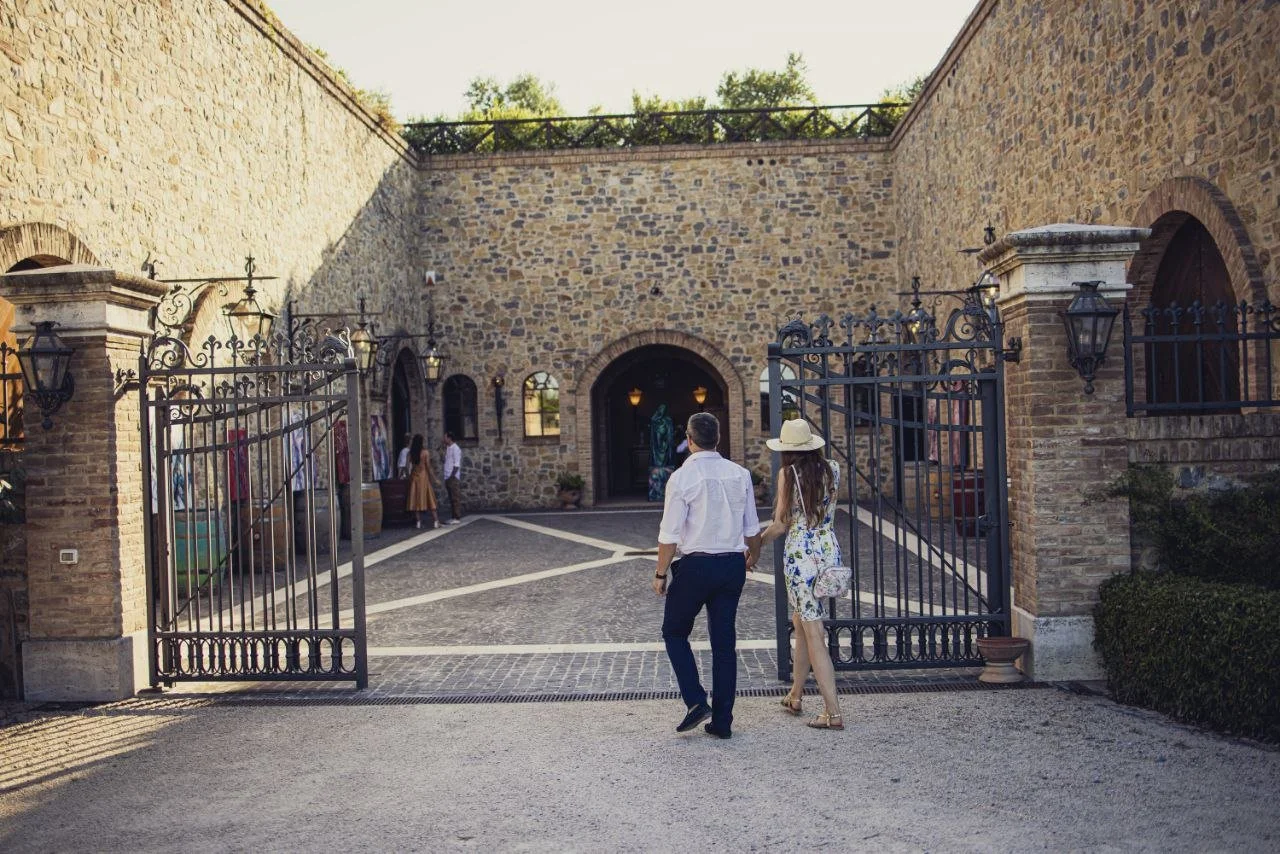 A couple entering a gated courtyard of a stone building, with people inside, in a sunny outdoor setting.