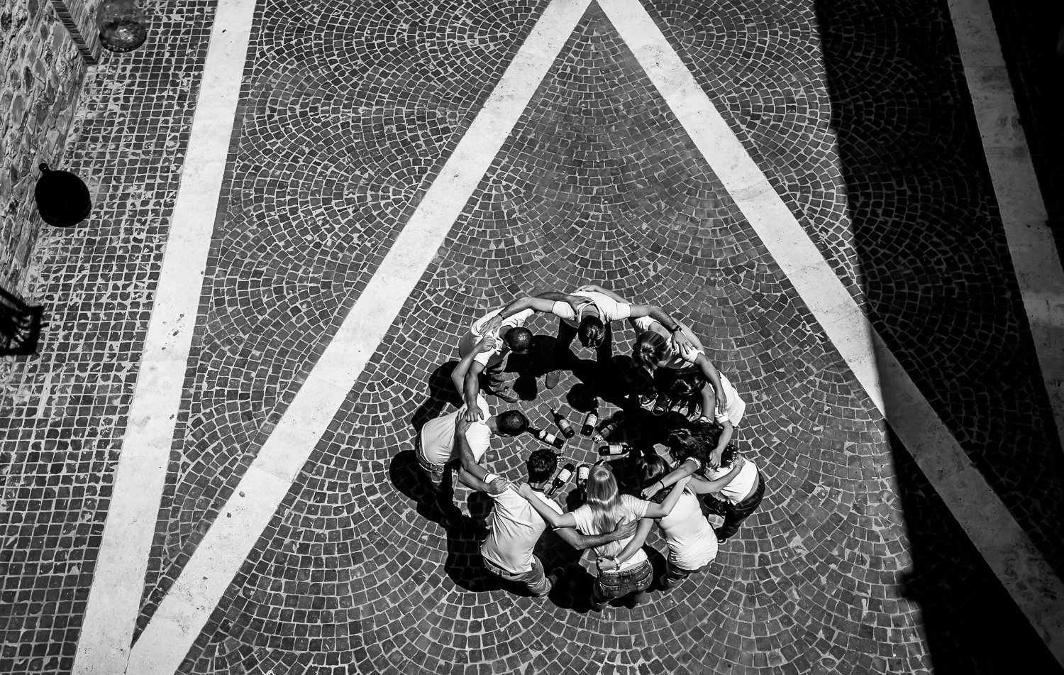 A group of people are standing in a circle on a cobblestone pavement, seen from above. They are engaging in a group activity with several bottles placed in the center.