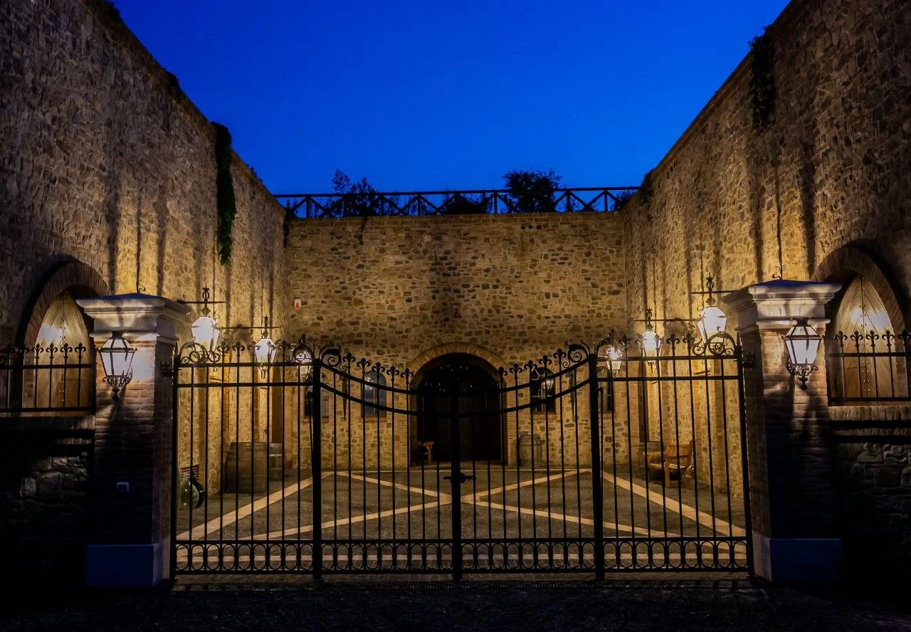 A gated courtyard with stone walls illuminated by wall-mounted lanterns during dusk, featuring a central archway and a blue evening sky.
