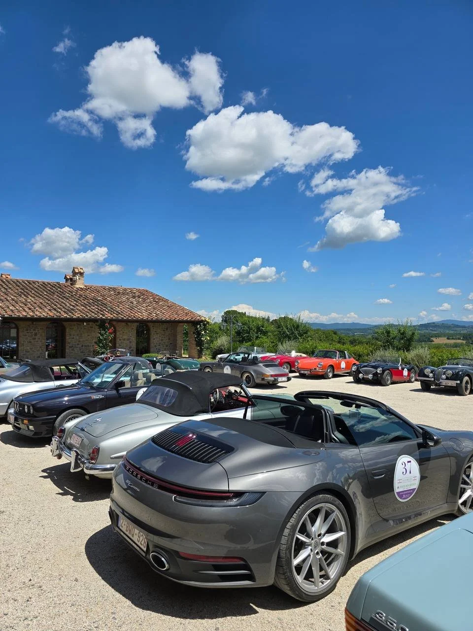 A collection of classic and modern cars parked outdoors on a sunny day with a clear blue sky and scattered clouds.
