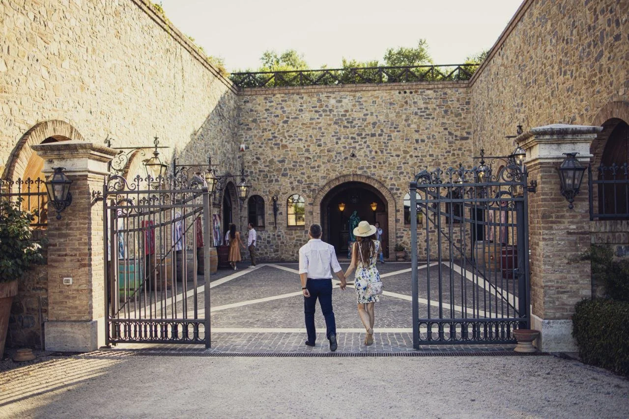 Couple holding hands walking through an open wrought iron gate into a stone courtyard with a stone building and art display on the sides.