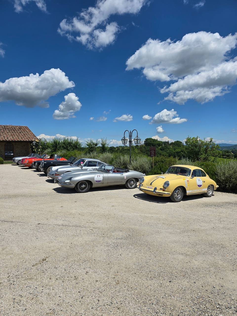 A lineup of vintage cars parked on a gravel lot under a blue sky with scattered white clouds, featuring a yellow Porsche and silver Corvettes, with greenery and a building in the background.