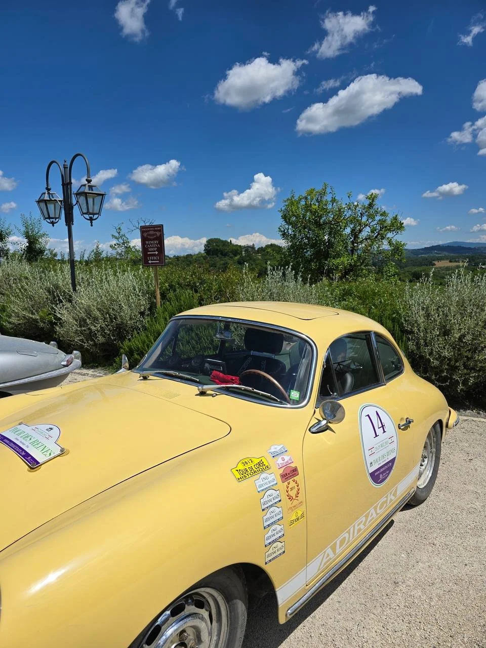 A vintage yellow race car with race stickers and a number 14 on the side, parked outdoors on a sunny day with greenery and a blue sky with white clouds in the background.