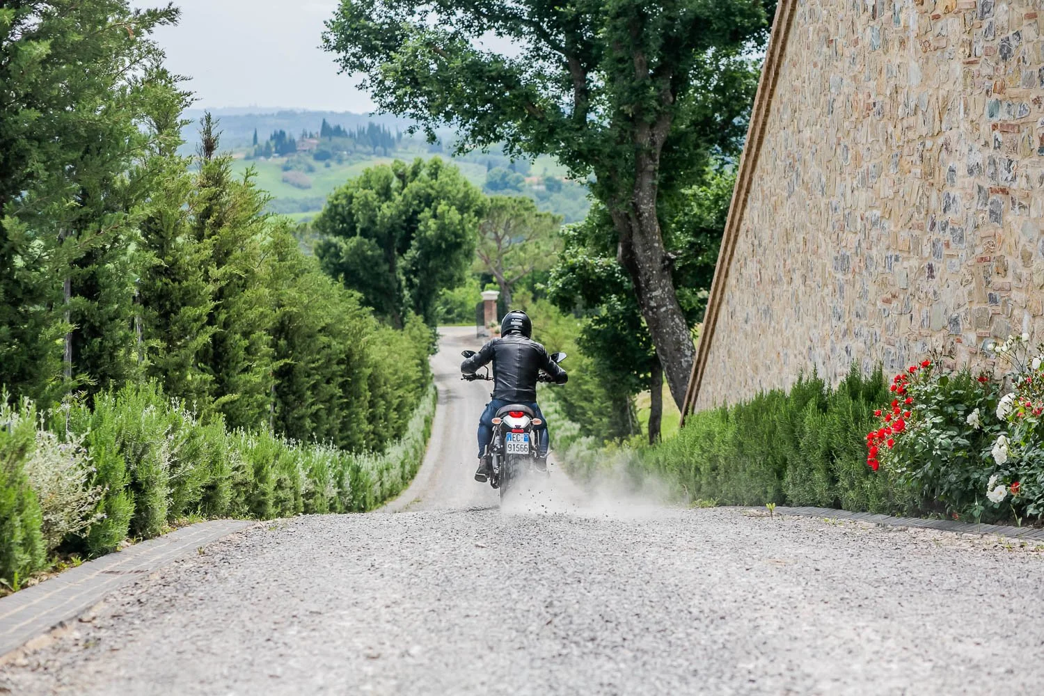 Motorcycle rider dressed in black riding on a gravel path through a lush green area with trees and bushes, with a stone building on the right and rolling hills in the background.
