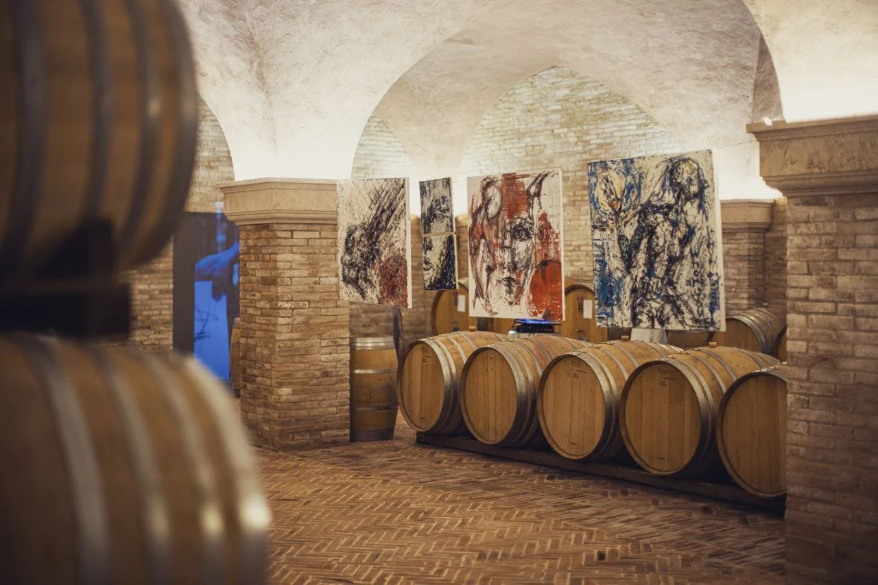 Art gallery in a wine cellar with paintings displayed on wooden barrels and brick walls.