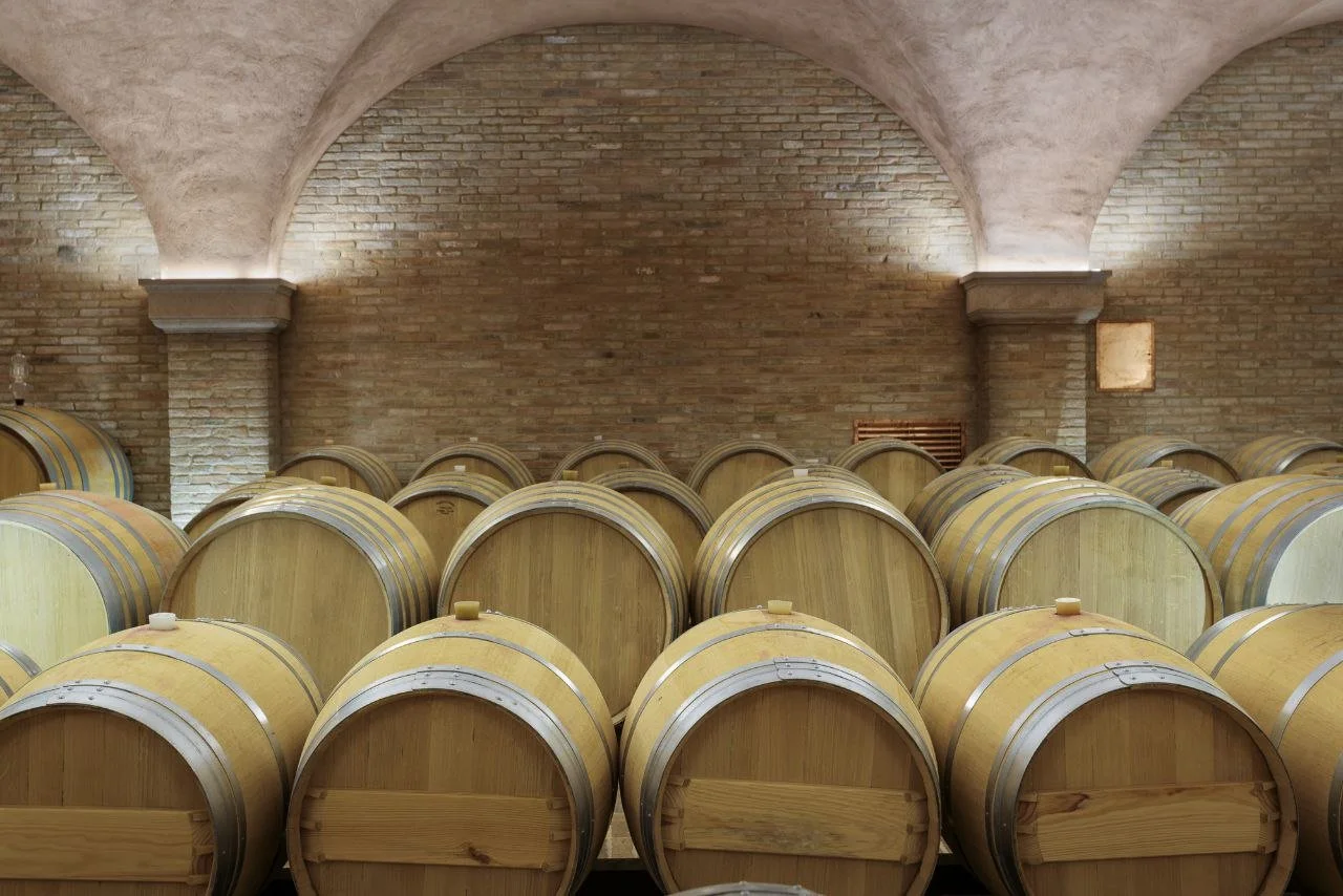Multiple wooden wine barrels stored in a cellar with brick walls and arched ceilings.