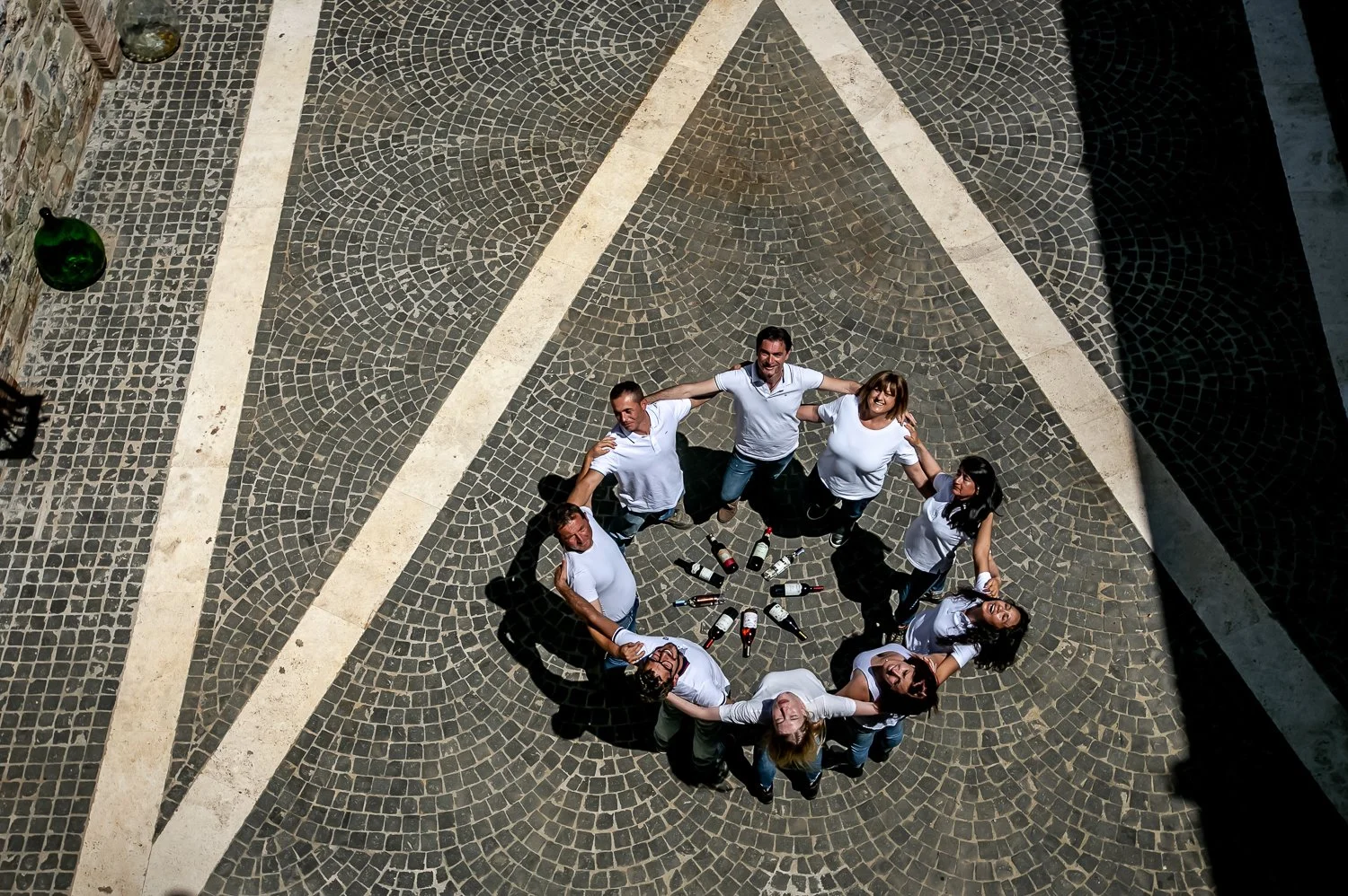 A group of seven people standing in a circle outdoors on a stone-paved area, holding each other’s shoulders, with empty beer bottles arranged in a circular pattern in the center.